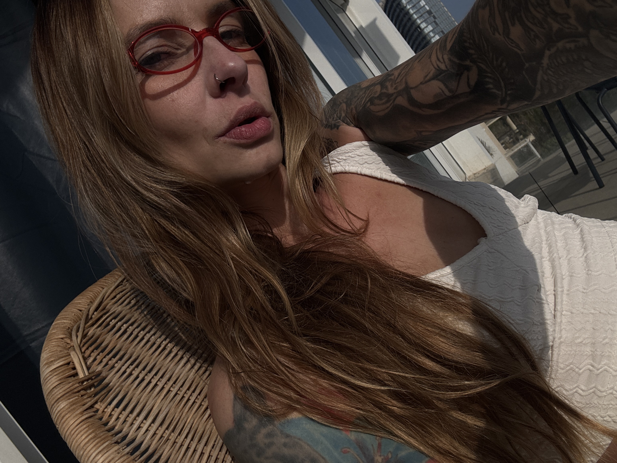 A woman with long brown hair, wearing red-framed glasses and a nose ring, is sitting in a wicker chair. She is wearing a white textured top. Her upper arm and shoulder have visible tattoos. The background shows a building with multiple floors and glass windows, suggestive of a city skyline. Light suggests an outdoor or sunlit environment. The general architectural style and climate suggest a location similar to Tel-Aviv, Israel.