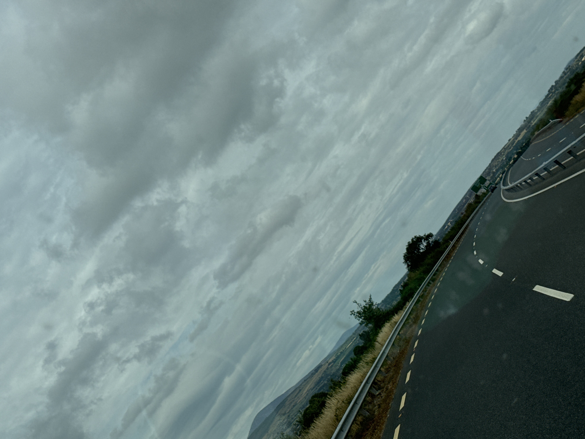 This is a tilted, eye-level, medium shot of a highway in the New Ross Municipal District, Ireland. The sky is overcast with thick, grey clouds that dominate the upper portion of the frame. A mountain range is visible in the distance on the left side of the image, partially obscured by the clouds. The highway is paved with asphalt and marked with white lines, including a dashed white line to the left of the lane and a solid white line to the right. The road curves to the right, with a guardrail separating it from a grassy embankment. Some dry grass and a few trees are visible on the embankment. The scene is likely captured from a moving vehicle, given the perspective and the slight blur in some areas. There are no people or animals visible in the image.