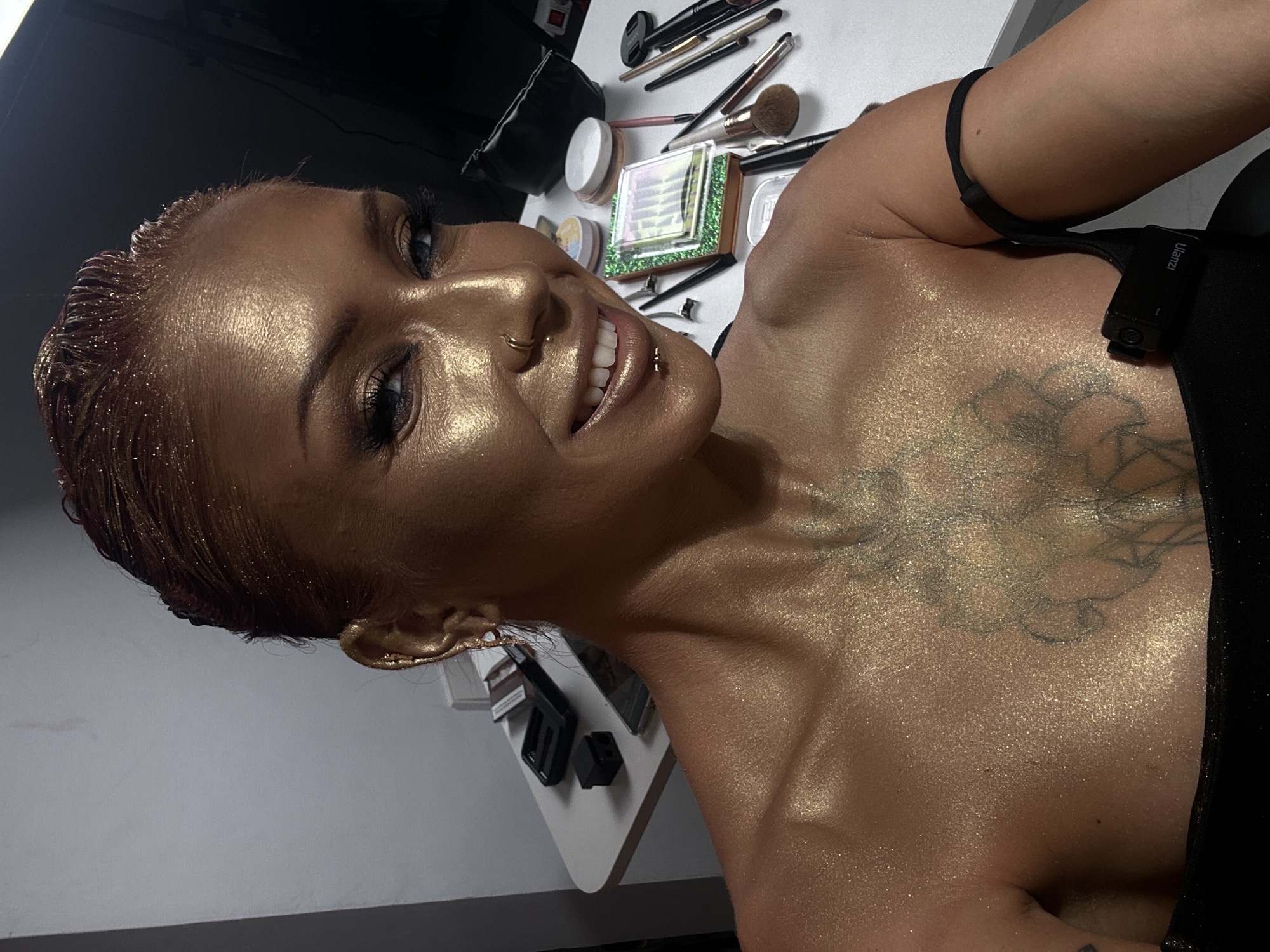 The media file captures a woman adorned with elaborate, shimmering golden body makeup. Her head is tilted, and she is smiling broadly, showcasing her white teeth. She has short, reddish-brown hair, which also appears to be dusted with golden glitter. Notable facial piercings include a gold ring in her nose and a gold stud on her lip. Her eyes are open, with defined eyebrows and visible mascara, contributing to a striking gaze.
Her entire visible skin, encompassing her face, neck, shoulders, and arm, is covered in a prominent, reflective golden glitter or body highlighter, creating a radiant and dazzling effect. A tattoo, appearing as a geometric or abstract design, is visible on her upper chest and shoulder area, partially gleaming beneath the golden shimmer. She is dressed in a black top or strap, to which a small black electronic device is clipped. The brand name "ulanzi" is distinctly visible on this device, suggesting it could be a wireless microphone transmitter or a camera accessory.
The background reveals a white surface laden with various makeup tools and products. These include multiple makeup brushes of different sizes, compact cases, pots, and jars. A distinctive square compact with green glittery edges and iridescent, stacked layers inside, possibly containing false eyelashes or an eyeshadow palette, is also visible. Hair clips are scattered among the items.
The scene suggests an indoor setting, likely a dressing room, studio, or backstage area where she has been prepared with professional makeup. Her confident smile and pose imply she is ready for a photoshoot, performance, or a special event in North Kuta, Indonesia.
