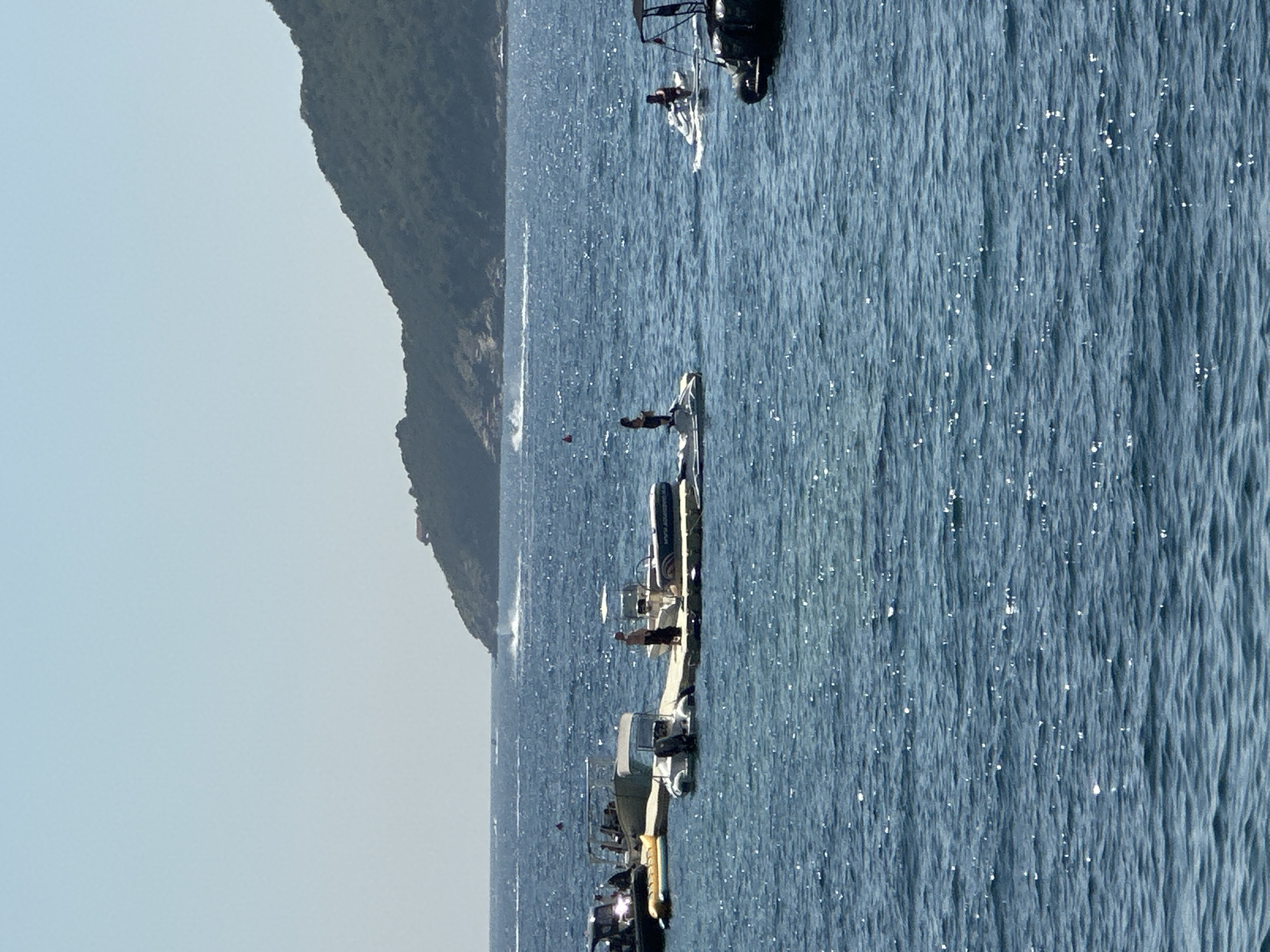 A sunny day at sea near Budva, Montenegro, features several boats and jet skis. In the foreground, a person stands on a floating dock with a few small boats tied to it. To the right, two jet skis are in motion, one with a rider and another appearing to be unoccupied. A larger boat with a canopy is visible on the far right. In the mid-ground, another floating dock supports more small boats and a few people. A yellow inflatable object is attached to a black boat on the left. In the background, a lush green hill slopes into the sea, with a building visible near the summit. The clear blue sky and sparkling water suggest a pleasant day for water activities.