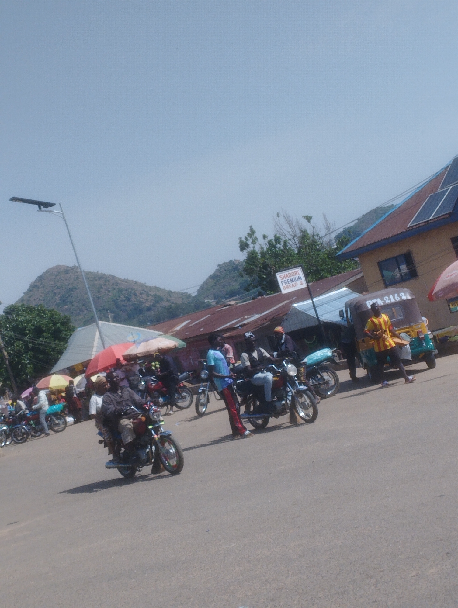 The image depicts a bustling street scene, likely in the city of Gwong, Nigeria, on a clear day. The foreground is dominated by a paved road with several motorcycles, some occupied by riders and passengers, and others parked. In the middle ground, a variety of market stalls are set up under colorful umbrellas, with people browsing and interacting. A distinctive three-wheeled auto-rickshaw is also present, with its license plate clearly visible as "PTA-2163". Behind the market stalls, buildings with corrugated metal roofs are visible, some equipped with solar panels. In the background, lush green hills rise against a pale blue sky, suggesting a location with natural beauty surrounding the urban area. A tall street lamp stands on the left side of the image. The overall impression is one of vibrant daily life and commerce within a semi-urban, possibly hilly, environment.