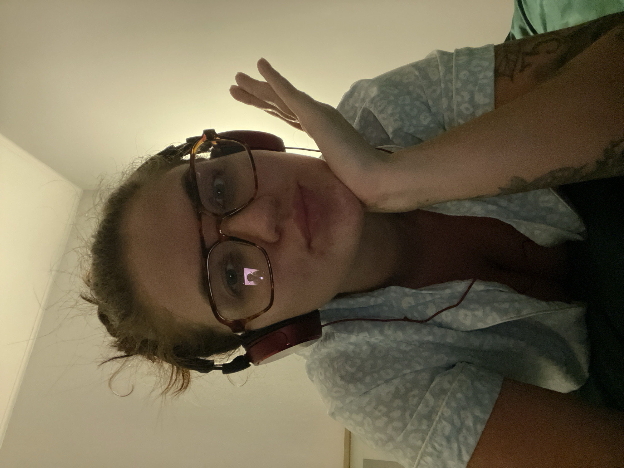 A woman with brown hair pulled back, wearing glasses and red headphones, is looking at the camera. She is wearing a light blue patterned shirt. Her right hand is raised near her face. She has tattoos on her left arm. The background appears to be an indoor space, possibly a room with a light-colored wall. No information or objects in the image can confirm the specific location in Мосметаллоконструкция Онт, Russia.