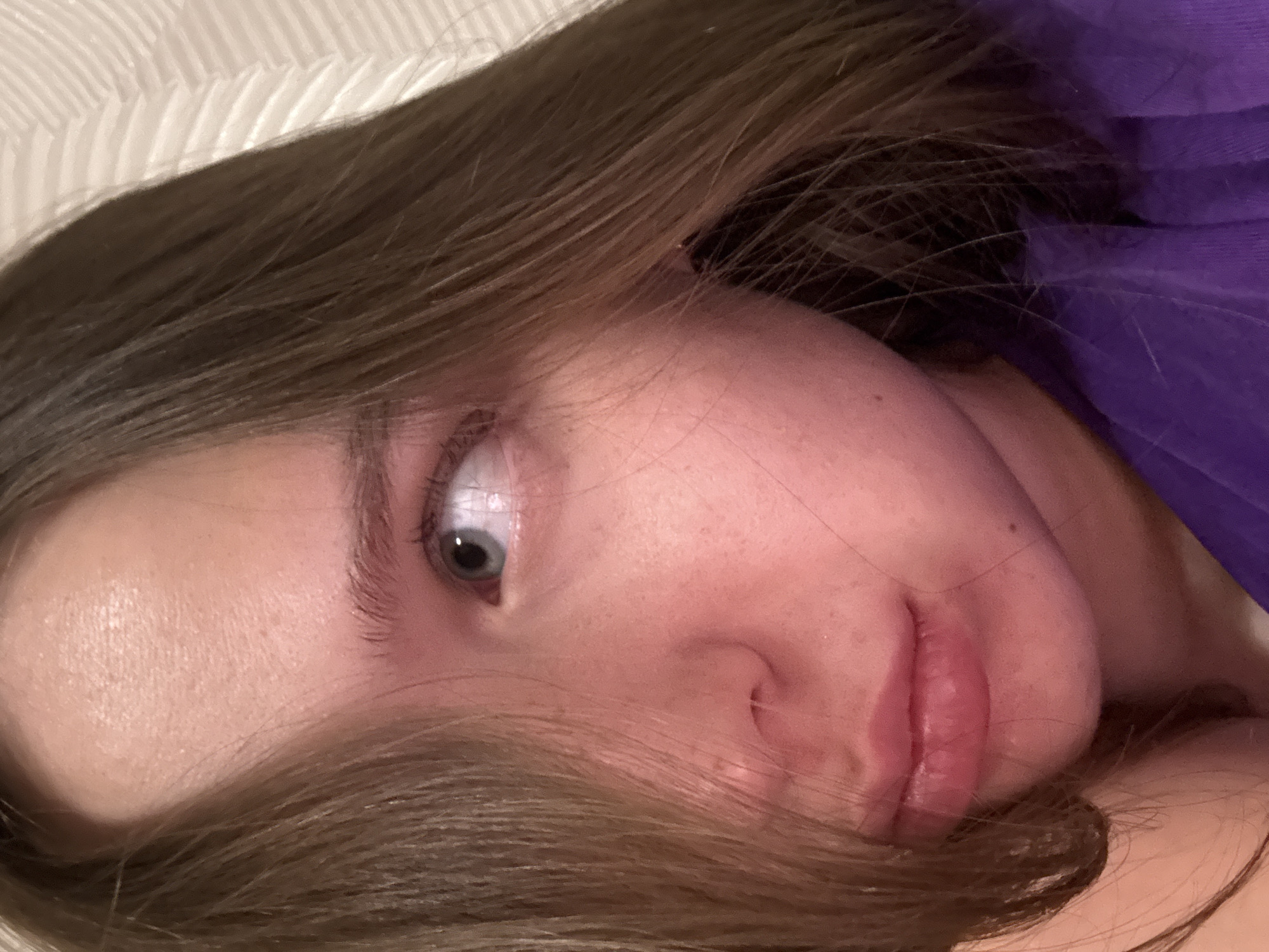 A close-up, indoor photograph shows a person lying down, angled so that their face is prominent in the frame. The individual has fair skin, light brown hair partially obscuring their face, and a single visible eye with a gray iris. The person is wearing a purple top. The background shows white bedding. The location in Chaikovsky, Russia, cannot be determined from the information provided in the image.