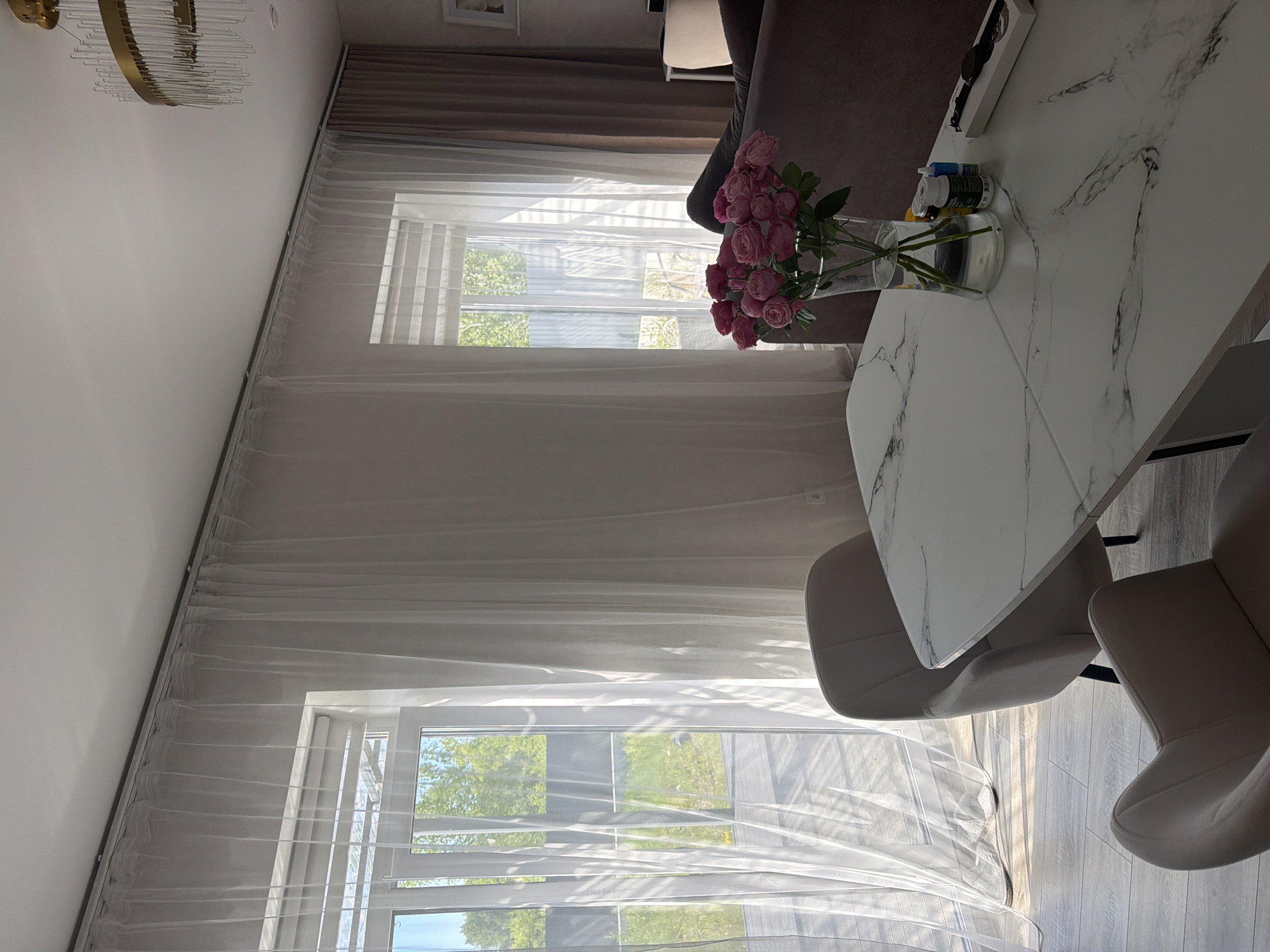 This is a photograph of a dining area in a residential setting, likely an apartment or house, in Malinovka, Russia. The scene is captured from a slightly elevated perspective, looking across a modern white marble-patterned dining table. On the table, a clear glass vase holds a bouquet of pink roses. To the right of the vase are several small bottles and a black watch in a white box. Two upholstered dining chairs, in a neutral beige color, are positioned at the table.
The background features large windows covered by sheer white curtains, allowing natural light to stream in. Through the windows, a glimpse of an outdoor area with greenery and a fence is visible. To the right of the sheer curtains, heavier, darker curtains are drawn. A glimpse of a dark-colored sofa is visible in the far right background, suggesting a living area adjacent to the dining space. A modern chandelier with glass accents hangs from the ceiling. The flooring appears to be light-colored wood laminate. The overall impression is one of a well-lit, contemporary interior space. There are no people visible in the image.