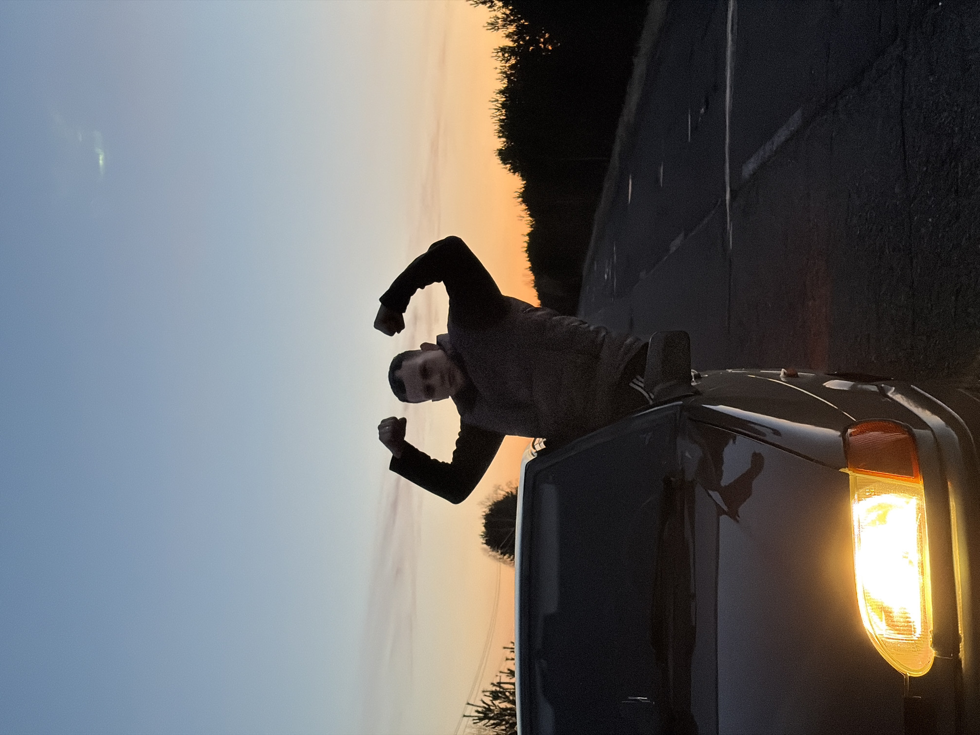 The image, originally rotated, captures an outdoor scene at dusk or dawn. A person, appearing male, is leaning out of the open roof or window of a dark-colored vehicle, positioned on the right side of the frame. The individual is looking towards the viewer with a slight grin or smile, flexing both biceps in a playful or proud pose. He is wearing a dark top, with parts of his silhouette visible against the bright sky.
The vehicle, possibly an SUV or truck, has its right headlight brightly illuminated, casting a warm yellow glow. The background features a vast, clear sky, displaying a gradient of colors that transition from a light blue on the left to warm orange and yellow hues on the right, indicative of either sunrise or sunset. Below the horizon, a dense line of dark trees or foliage creates a stark, silhouetted edge, running alongside what appears to be a road with subtle markings.
The overall lighting is dim, dominated by the natural light from the sky and the artificial light of the car's headlamp. No specific weather conditions are discernible beyond a clear sky. There is no visible text in the image. The scene suggests a stop along a road, potentially in a rural area, with the contextual location being Kharnavy, Russia.