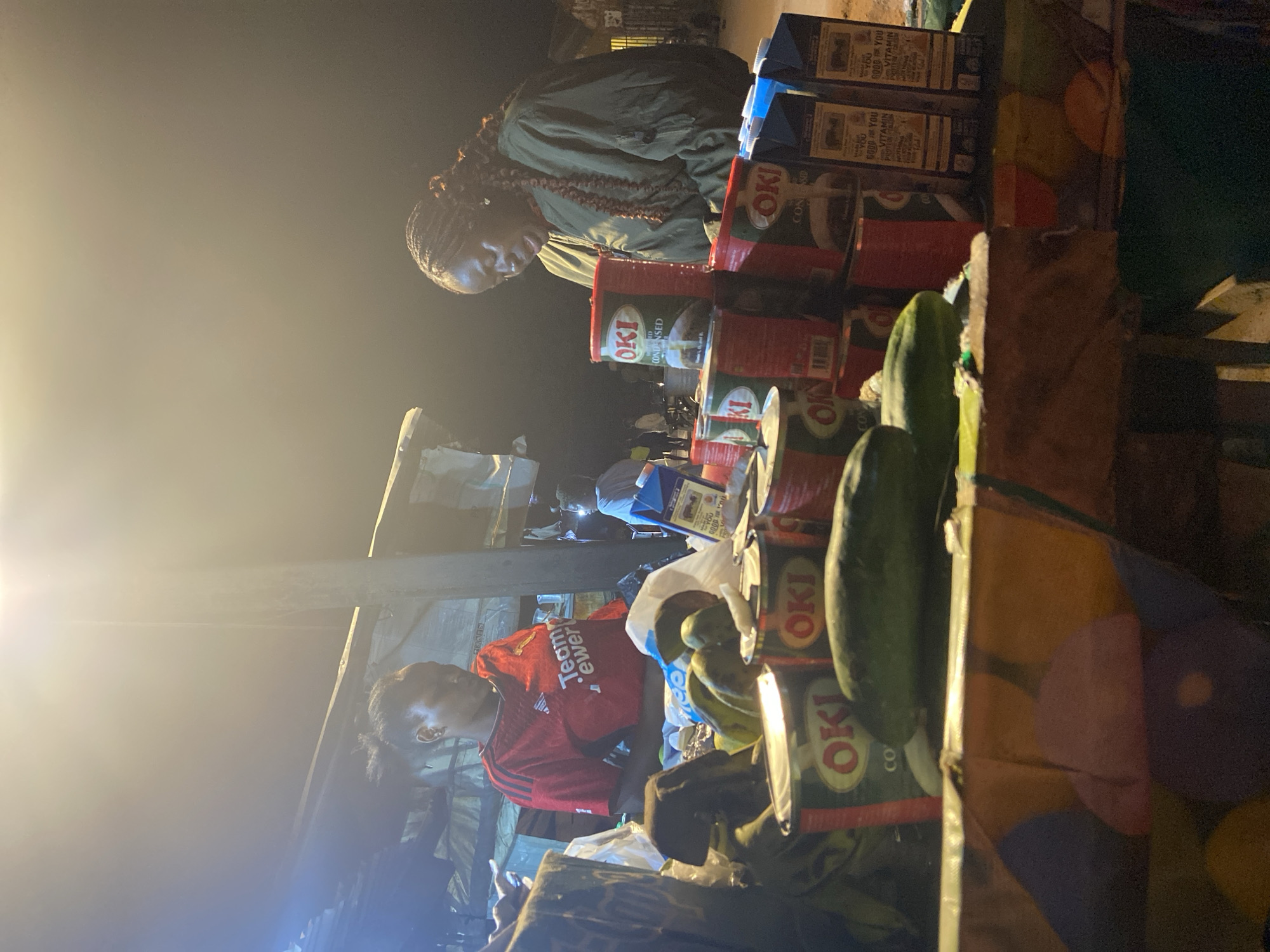The image depicts a nighttime market scene in Du, Nigeria. Two women are visible, seemingly engaged in a transaction or conversation. The woman on the left, wearing a red t-shirt with "Teamewer" visible, stands behind a display of canned goods and produce. The woman on the right, wearing a green jacket, is looking down at the merchandise. The foreground of the image is dominated by stacks of "OKI CONDENSED MILK" cans and some vegetables, including cucumbers. In the background, more market stalls and structures are dimly lit. The overall atmosphere suggests a bustling local marketplace after dark.