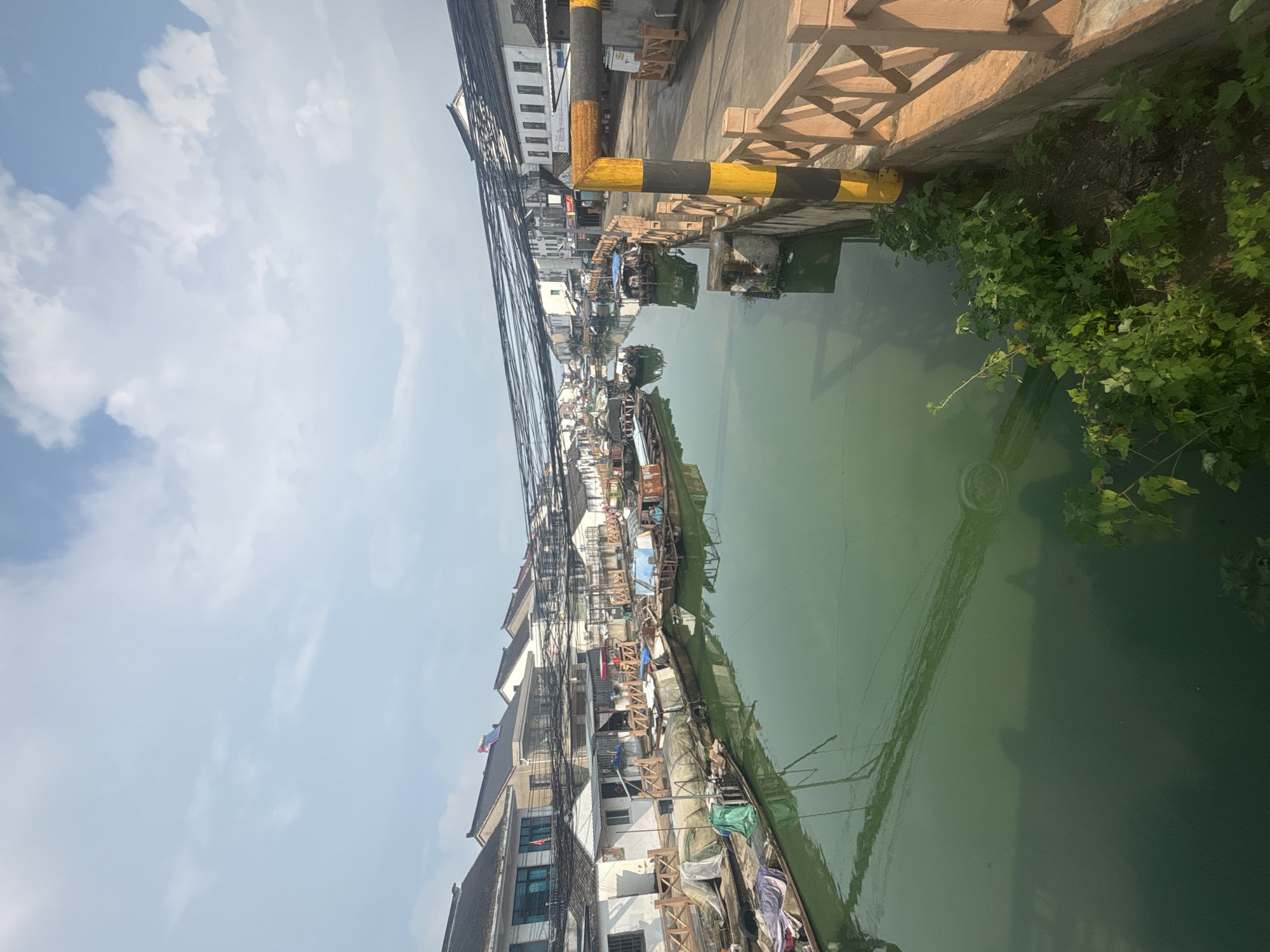 This is a vertical, eye-level, outdoor shot of a canal in Wuzhong District, China. The canal is lined with traditional Chinese architecture on both sides, featuring whitewashed walls and dark tiled roofs. Several boats are moored along the canal, some covered with tarps, others appearing to be residential or for transport. The water in the canal is a murky green. Overhead, a dense network of black wires crisscrosses the sky above the buildings and boats. On the right side of the frame, a wooden railing runs along the edge of a walkway, with a black and yellow striped pole in the foreground. Lush green vegetation grows along the bank near the railing. The sky is a pale blue with scattered white clouds.