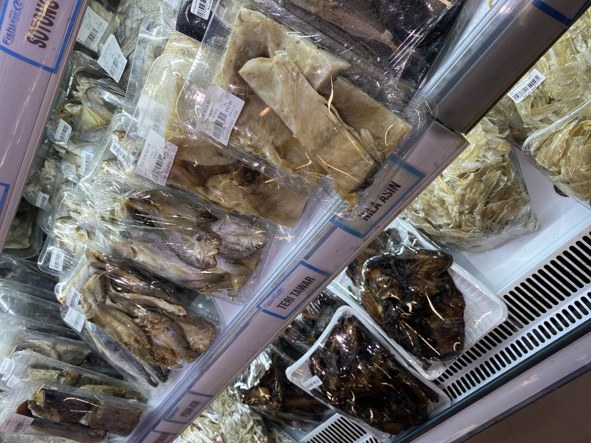 The image shows a retail display of dried fish products in plastic packaging, arranged on refrigerated shelves. Several labels are visible, including "Fish & Meat," "SOTONG," "TERI TAWAR," and "NILA ASIN." One label indicates the product is from "SUKUN MALI Banda Aceh." The dried fish vary in size, color, and form, with some appearing whole and others filleted or shredded. The scene is indoors, likely a supermarket or market stall in Banda Aceh, Indonesia, given the labeling. There are no people visible in the image.