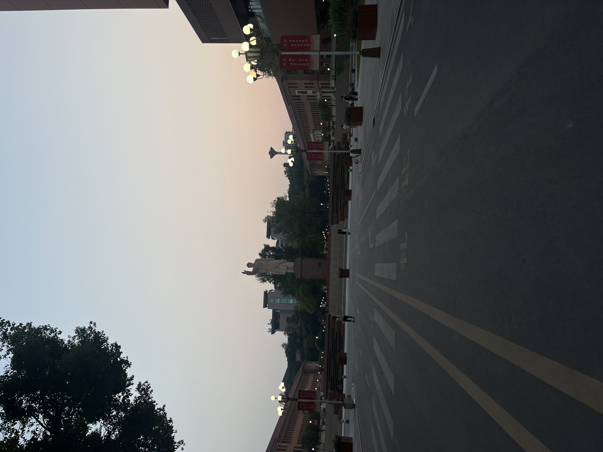 The image depicts a wide outdoor scene within a large institutional or university campus in Yuzhong District, China, observed at dusk. The sky transitions from a light blue overhead to a faint orange-pink hue near the horizon.
In the foreground, a dark asphalt road occupies the lower portion of the frame. It features white zebra crosswalk markings and double solid yellow lines down its center. Faded yellow Chinese characters are painted on the asphalt in the right lane.
The midground is dominated by architectural structures and landscaping. A prominent, light-colored statue of a male figure with a raised right arm stands on a tall, rectangular pedestal in the center-left. To the left of the road, a long, multi-story building with a brick-red facade and numerous rectangular windows is visible. To the right, a section of a modern, multi-story building with a dark brown/red exterior and complex geometric patterns is partially seen. Behind the central statue, several modern buildings with glass elements are situated on a tree-covered hillside.
The area features extensive landscaping, including wide, dark-colored steps or terraces leading up towards the buildings and statue. Rectangular red planters containing green foliage are positioned along the road and steps. Numerous ornate street lamps, each equipped with multiple spherical white light fixtures, are illuminated, casting a warm glow. Red banners with white Chinese characters hang from several of these street lamps, particularly on the right side of the road.
Several individuals are present, appearing as dark silhouettes against the ambient light. One person walks across the crosswalk in the center, moving from left to right. Other figures are visible near the street lamps and in the distance by the buildings.
The background shows a densely forested hill with additional structures nestled among the trees. A large, dark tree branch with dense foliage partially obscures the upper-left corner of the image.