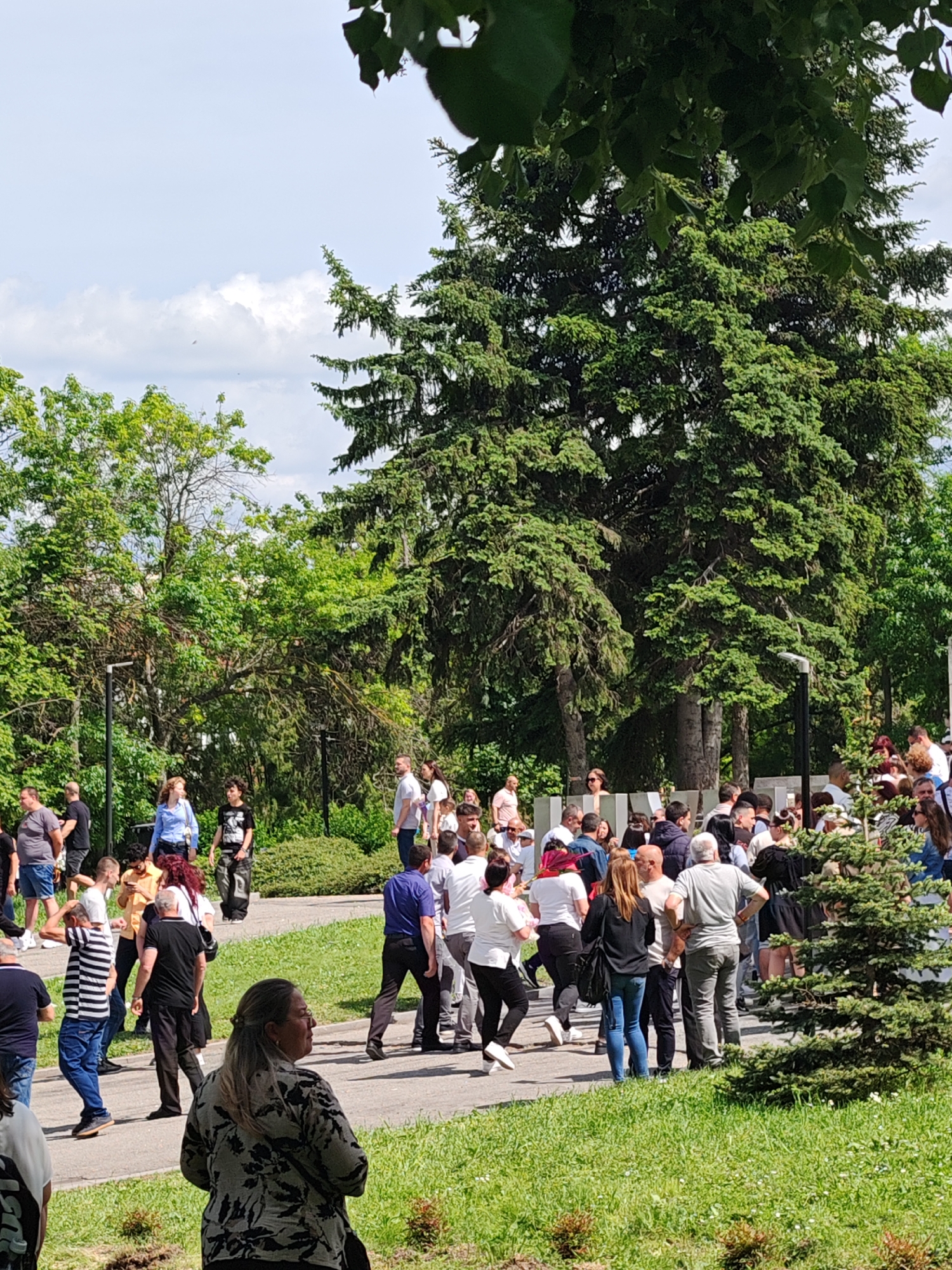 The image shows a large group of people gathered on a paved pathway in what appears to be a park. The location is the city of Shumen, Bulgaria. The pathway is surrounded by grass and various trees, including a large evergreen tree. Many of the people are dressed casually in a mix of light and dark clothing. There are more adults than children, and they are engaged in various activities such as talking, walking, and standing in groups. In the background, there are more trees and the sky, which has some clouds.