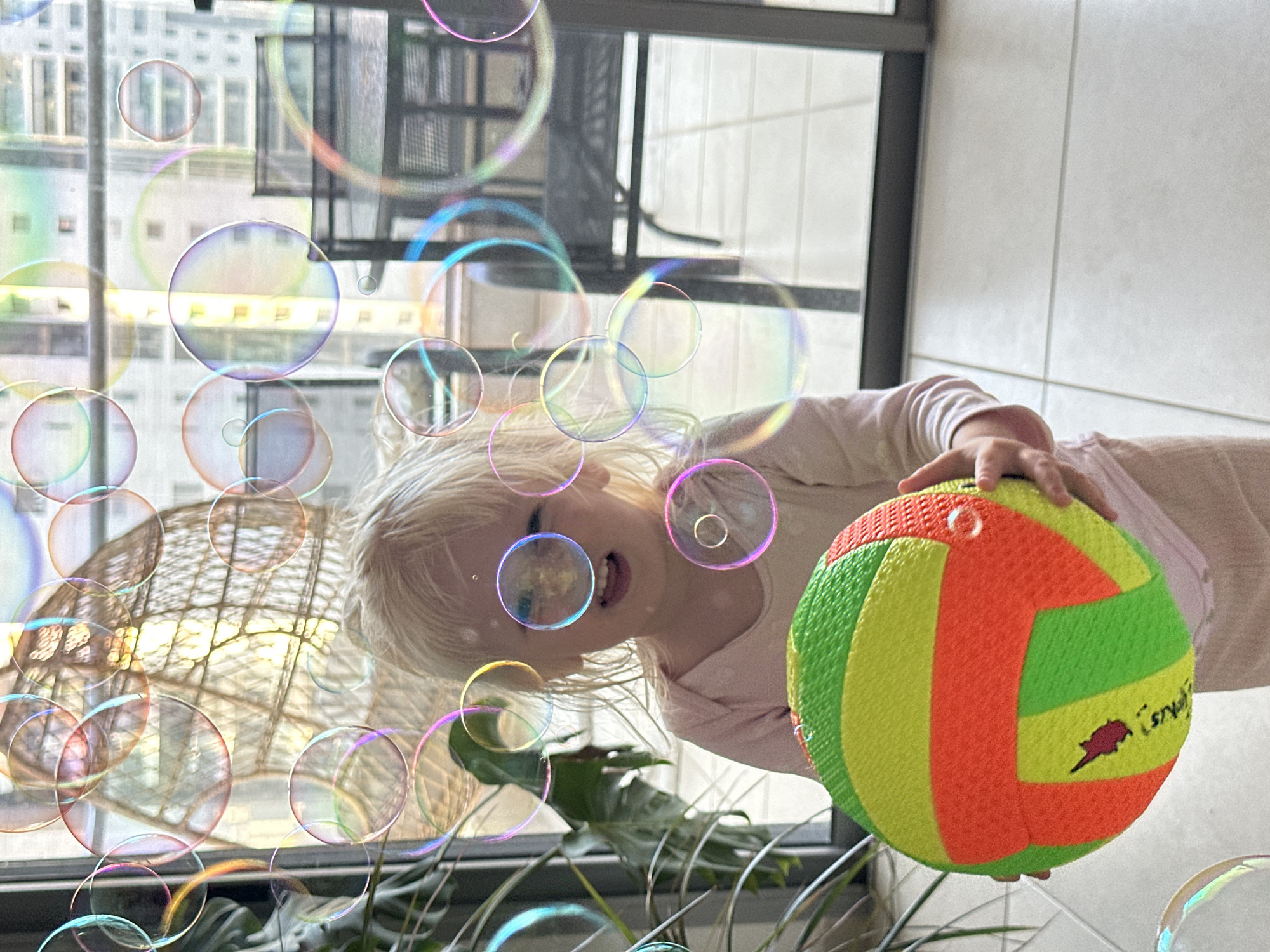 A young child with blonde hair, wearing a light pink long-sleeved shirt and light pink pants, stands indoors near a large window. The child is holding a colorful, textured ball with green, yellow, and orange panels. Numerous iridescent soap bubbles float in the air around the child and in front of the window. Through the window, a woven hanging chair is visible, as well as outdoor furniture and buildings in the distance, suggesting an urban environment in Petah Tikva, Israel. The floor is tiled. The scene appears to be a playful moment, possibly in a home.