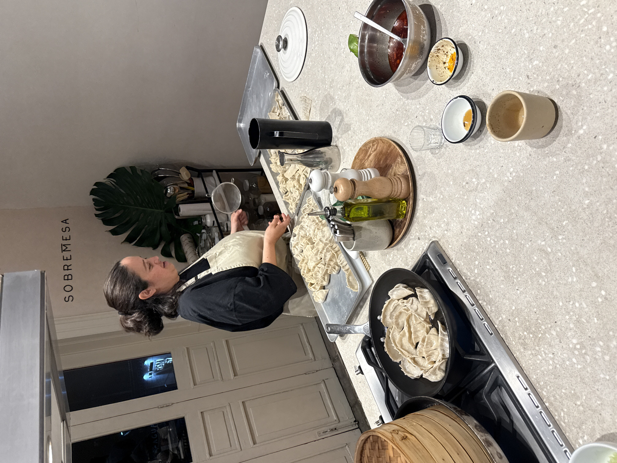 A woman is cooking at a kitchen counter. The image appears to be shot indoors in the city of Mexico City, Mexico, as indicated by the writing "SOBREMESA" which is a brand known to be based in Mexico City. The woman has dark hair pulled back in a bun, wearing a black shirt and an apron. She is working with food, possibly making dumplings or pasta, and there is a pan of food on a stovetop next to her. Other kitchen items are visible, including a bowl of ingredients, a steamer, utensils, and various containers. The overall scene suggests a cooking or food preparation environment.