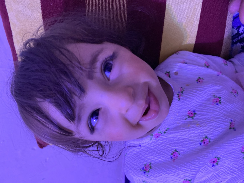 A close-up portrait shows a young child, likely a toddler, in an indoor setting within Kandahar, Afghanistan, under prominent purple illumination. The child has dark brown hair with bangs and appears to have kohl applied to the lash lines of her wide, dark eyes. Her fair complexion shows blush on the cheeks. The child displays a playful expression, with her mouth slightly open and tongue partially extended, revealing some upper and lower teeth. She is wearing a light-colored, long-sleeved top with a textured surface, patterned with small pink, purple, and green floral motifs. Minor light brown stains are visible on the front of the garment. To the child's right, a cushion or pillow featuring alternating horizontal stripes of dark maroon and light yellow is present. A small fragment of blue and white patterned fabric is visible at the bottom right. The background to the child's left is a plain, textured surface.