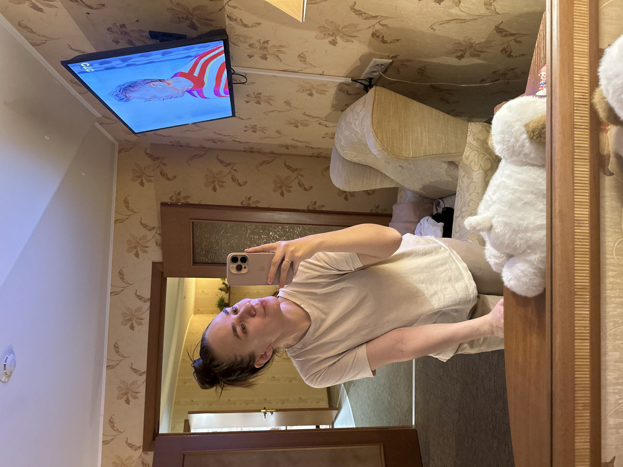 A person is taking a selfie in front of a mirror. The person has their hair pulled back in a bun and is wearing a light-colored t-shirt. The background shows an interior space with patterned wallpaper and a TV mounted on the wall. The TV is displaying a person wearing a striped shirt. There is a beige chair and a white stuffed animal visible near the mirror. The location appears to be a room inside a home. The city and country cannot be determined from the image.