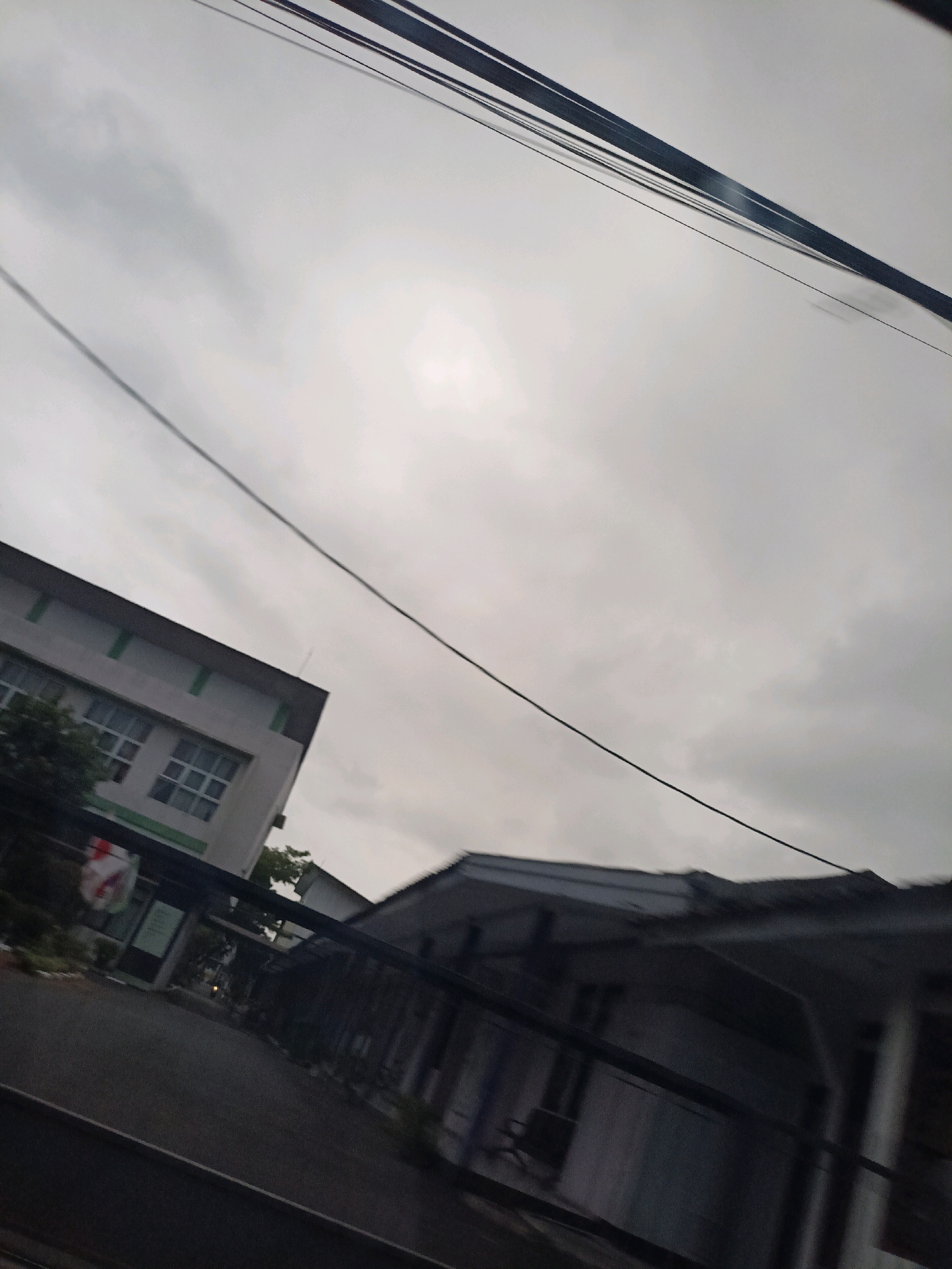 The image depicts an outdoor scene, likely a street view, with a building on the left and another structure in the foreground. The sky is overcast. Several wires are visible, running diagonally across the frame. The building on the left appears to be a multi-story structure with windows. The foreground structure has a dark, sloped roof and appears to be a building or shelter. The overall impression is one of a cloudy day in an urban or semi-urban environment. The specific location in Magelang, Indonesia cannot be determined from the visual information alone.