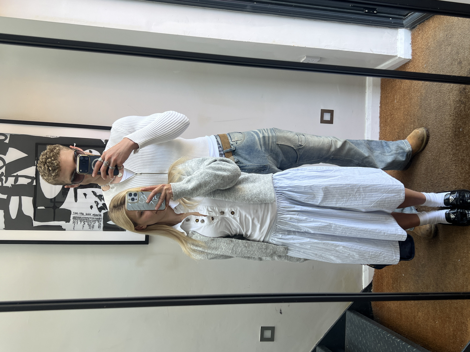 A vertical, medium shot captures a couple posing in front of a mirror. The man, on the left, has short curly hair and wears sunglasses, a white ribbed zip-up sweater, light wash jeans, and a brown belt. He holds a black smartphone in his right hand to take the picture. The woman stands to the right of the man, wearing a white button-down dress, a gray cardigan, white socks, and black loafers. She also holds a smartphone. The background includes a white wall, a piece of modern art framed in black, a dark-colored window frame, and a section of what appears to be a staircase. The setting is indoors, with a carpeted floor visible. Based on architectural details, it is possible the location is a residential or commercial building in London, United Kingdom, but this could only be confirmed through additional information.