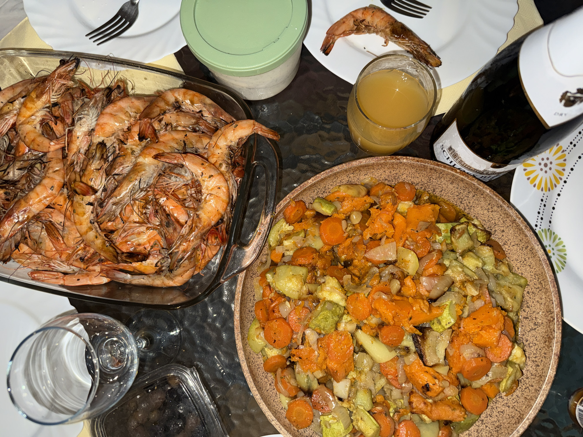 The image presents an overhead view of a dinner table laden with a festive meal, likely set indoors during the evening in Bar, Montenegro. The table surface is dark and somewhat reflective.
Two main dishes dominate the scene. On the left, a large, clear rectangular glass dish is piled high with numerous cooked shrimp or prawns. These are primarily reddish-orange, many with heads and shells intact, and appear to be seasoned. To the right, a large, round, rustic brown bowl holds a colorful medley of cooked vegetables, featuring bright orange sliced carrots, light green chopped zucchini or similar squash, and translucent white onions.
Various drinks and tableware complement the meal. A clear glass contains a golden-orange liquid, possibly juice or a beverage. Next to it, a dark-colored bottle, likely wine, stands with a partially visible white label bearing the text "SCHLOSS". White plates and silver forks are scattered across the table; one plate holds a single cooked shrimp. A light green lidded container, possibly holding a sauce or dip, is placed near the shrimp dish. An empty clear wine glass is visible in the bottom left, along with a small, partially seen clear plastic container. A decorative plate with a yellow and black floral pattern is also visible on the bottom right. The artificial lighting suggests an indoor setting. No people are present in the image.