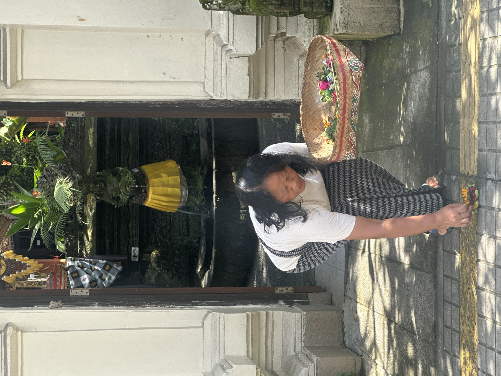 A woman wearing a white t-shirt and a black and white patterned skirt kneels on a paved sidewalk in front of an open doorway. She is placing a colorful offering of flowers on the ground from a woven basket held in her right hand. The doorway leads to an interior courtyard with a stone fountain and lush green plants. The building is constructed of white stone with a dark wooden doorframe. The scene is bathed in sunlight, casting shadows on the pavement. The location is identified as Ubud District, Indonesia, a region known for its spiritual significance and vibrant cultural traditions.