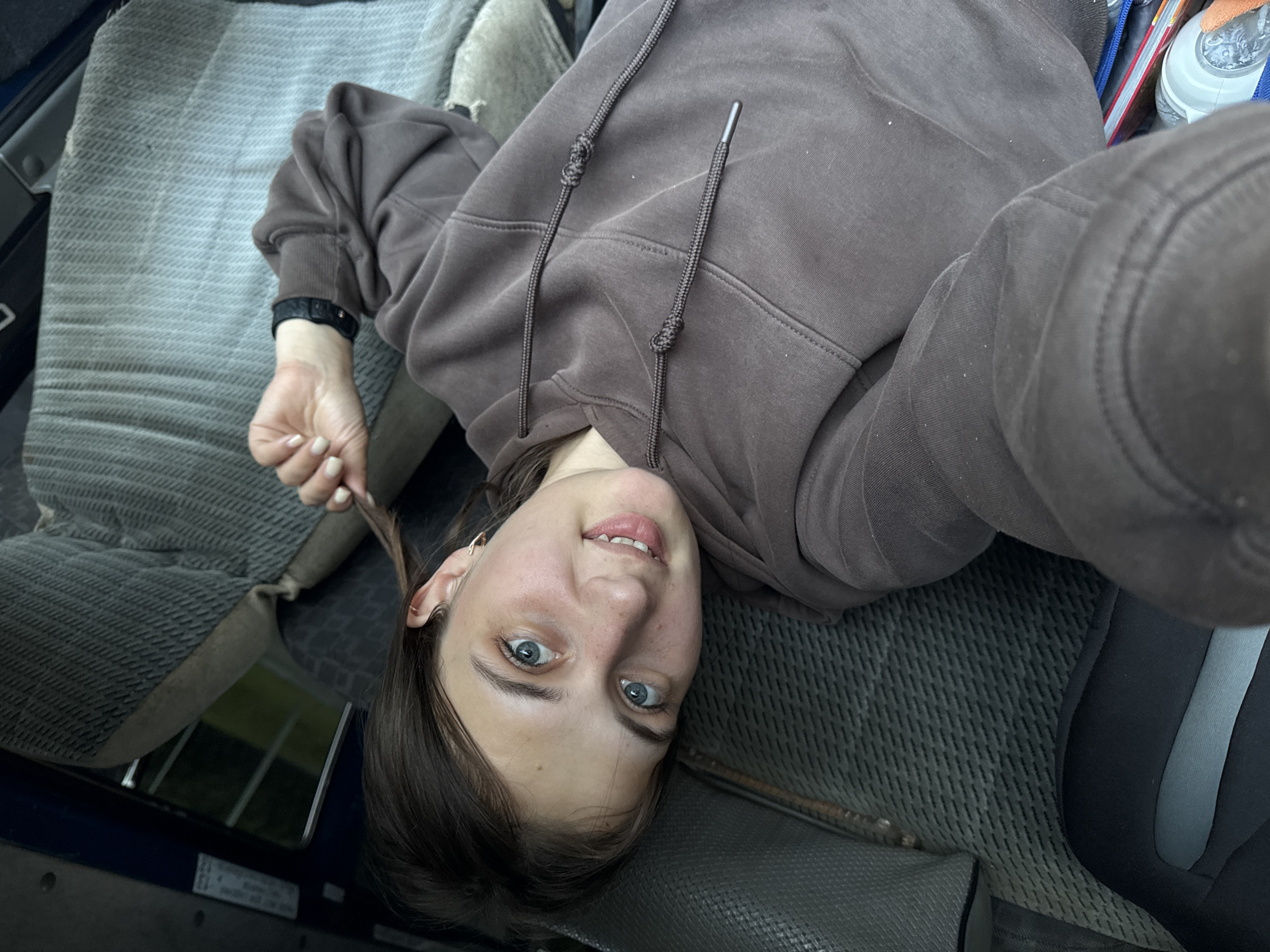 A young woman with blue eyes and brown hair is lying on her side, looking at the camera. She is wearing a brown hoodie and has a watch on her left wrist. Her right hand is near her face, holding a strand of her hair. She is in what appears to be the interior of a vehicle, possibly a van or truck, with worn, textured grey seats visible. The lighting suggests it is daytime, and the scene is likely outdoors given the glimpse of green foliage through a window. There is no clear indication of the specific location in Vary, Russia.