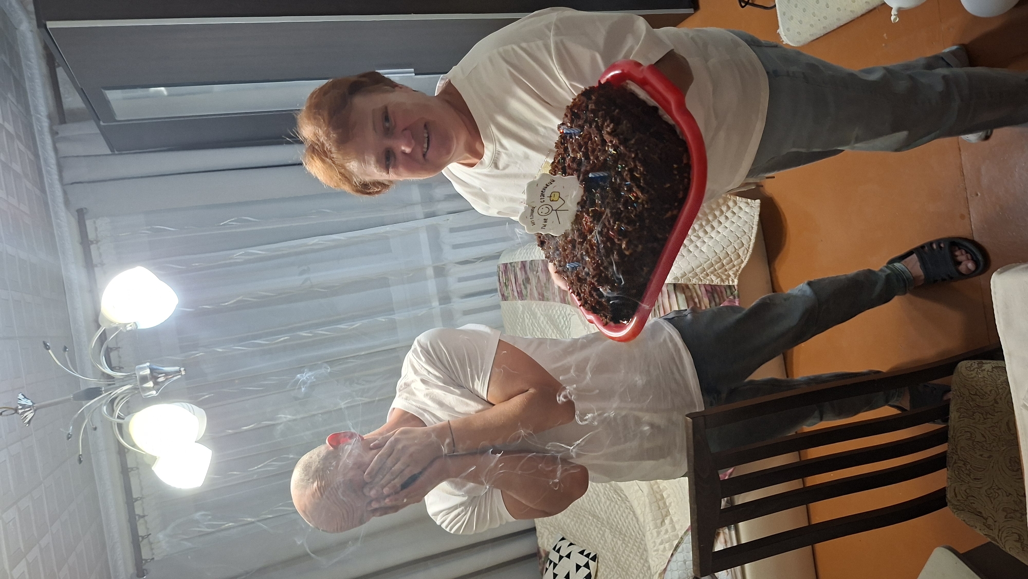 A mid-shot photograph, taken indoors, captures a celebratory moment. A woman with reddish-blonde hair and a warm smile holds a dark, frosted cake with lit candles and a small decorative element on top. She is wearing a white t-shirt and light-colored jeans. To her left, a bald man is seen from the shoulders up, with his hands covering his face, and smoke or vapor emanating from his shirt. He is wearing a white t-shirt and jeans, with dark sandals on his feet. The background reveals a room with a ceiling light fixture, light-colored curtains, and a sofa. The floor appears to be a dark, orange-brown hue. A chair with a patterned cushion is visible in the foreground on the left. The overall impression is of a birthday or special occasion. The image is dated July 27, 2023.