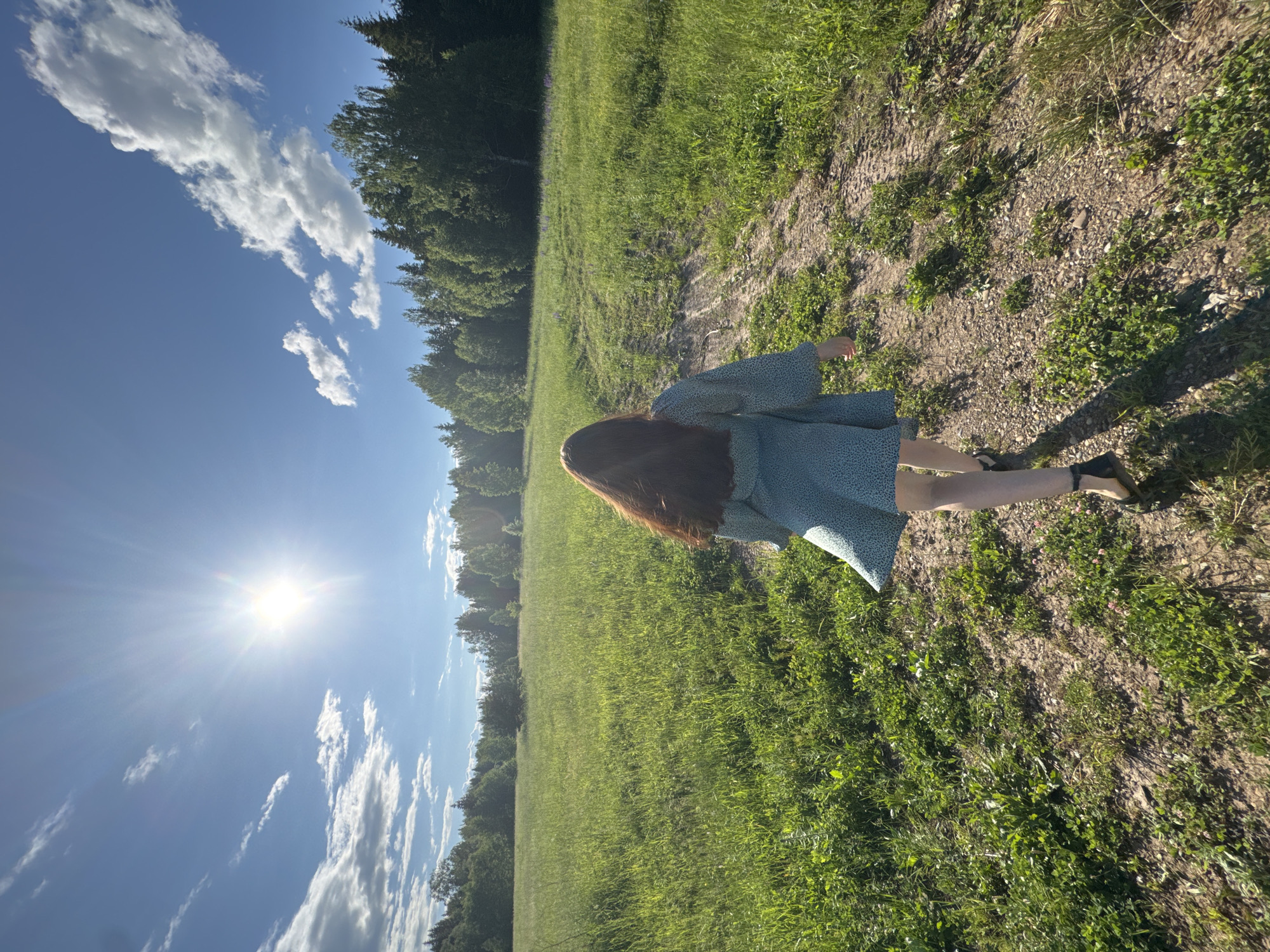 The image shows a young woman with long, dark hair walking away from the camera on a dirt path in a grassy field. She is wearing a light blue, patterned dress and dark sandals. The sun is shining brightly in a blue sky with scattered white clouds. A line of trees borders the field in the distance. The location is identified as Sosnovo, Russia.