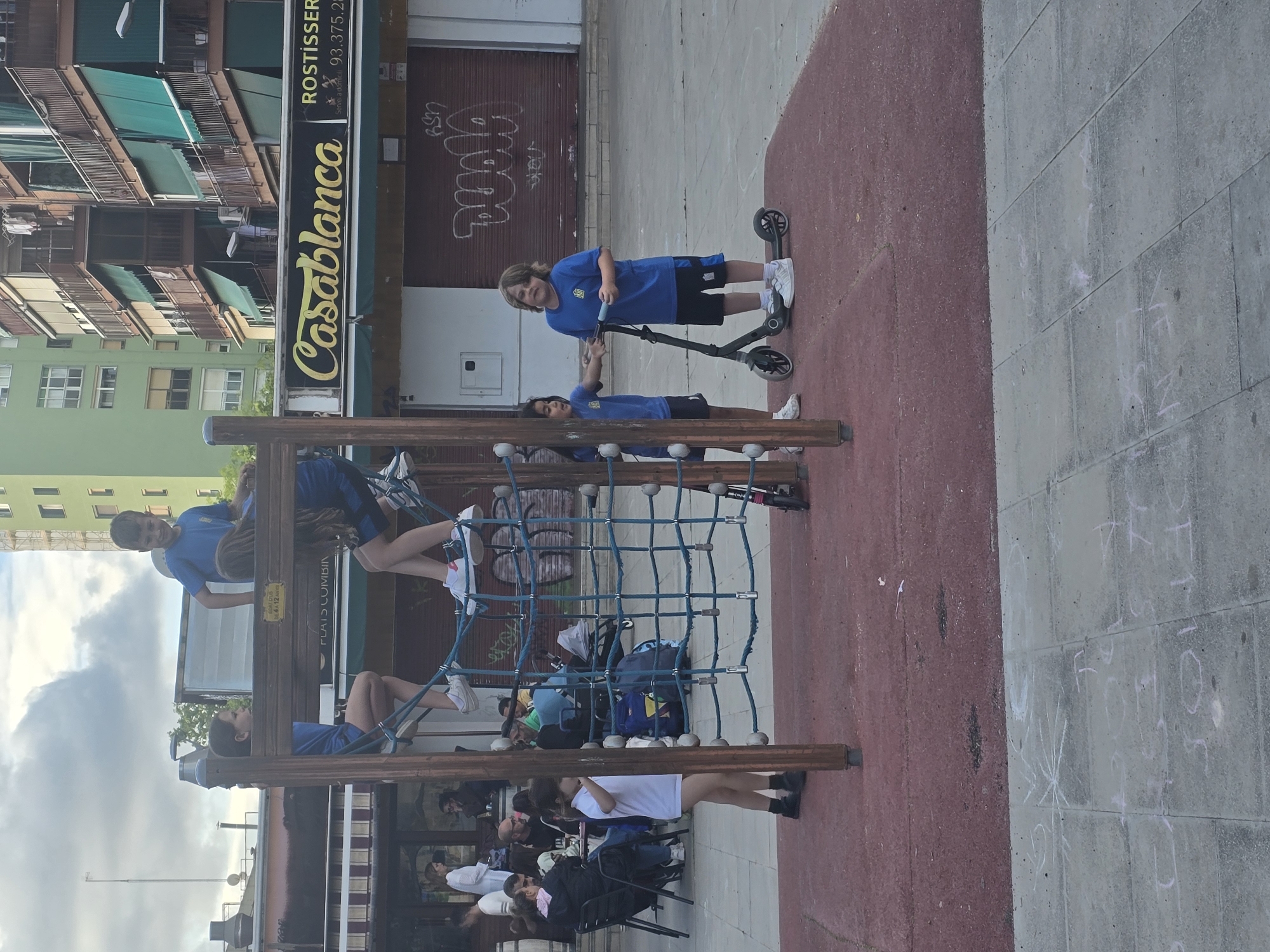 The image captures a scene in Cornellà de Llobregat, Spain, featuring a children's playground area with a climbing structure and a gathering of people seated at tables nearby.
Several children wearing matching blue and black athletic outfits are actively using the climbing structure. Two boys are riding a scooter close to the playground area.
A group of individuals is seated at tables next to the playground, likely patrons of a restaurant or café.
Behind the playground is a commercial building with a sign that reads "Casablanca." The architectural style suggests a residential area within the city.
The ground around the playground is a combination of red flooring and grey concrete. The weather appears fair, with partial cloud cover in the sky.