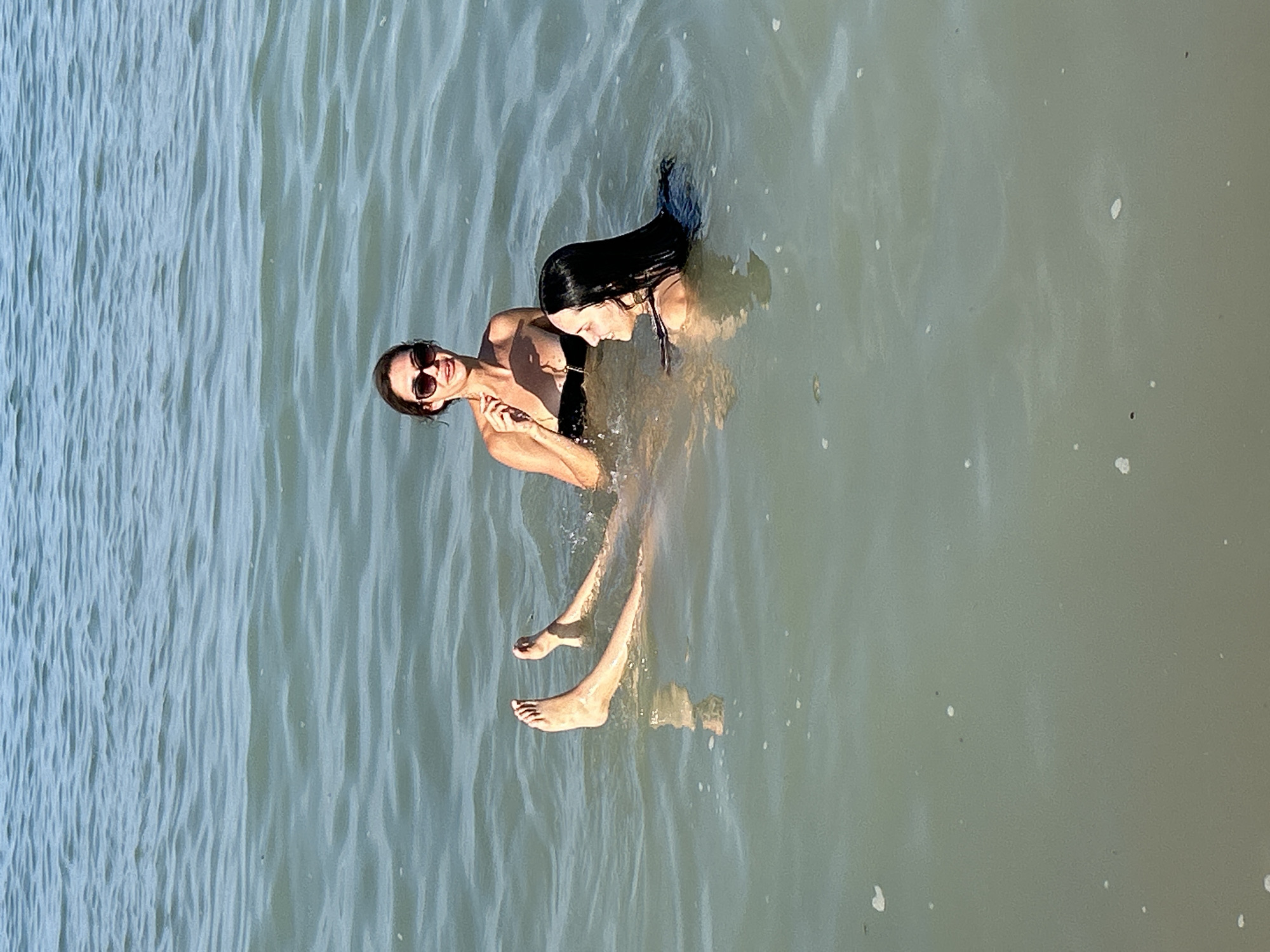 Two individuals are swimming in the water. One person, wearing sunglasses and a bikini top, is positioned on the left side of the frame, looking towards the camera with a smile. Their arms are clasped in front of their chest. The second person, with long dark hair, is behind and to the right of the first person, facing away from the camera and looking down. Both individuals appear to be enjoying themselves. Their lower bodies and legs are submerged in the water, with some splashing visible around their feet. The water is calm with visible ripples and reflections. The scene suggests a sunny day, likely in a natural body of water like a lake or the sea, given the light and the texture of the water. There are small specks of debris or foam on the water's surface. The image orientation is vertical, with the subjects oriented horizontally within the frame.