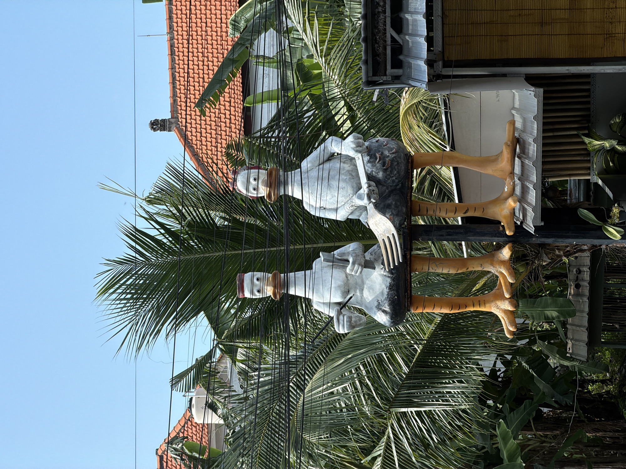 The image shows a whimsical outdoor sign in Kuta, Indonesia. The sign features two identical, cartoonish statues of a white bird-like figure with a red hat, clutching a stone-like object and a large fork. The figures' legs are painted in a striped yellow and black pattern, ending in large, orange feet. The sign is positioned amongst lush green palm trees and other tropical foliage. Above the sign, there's a building with a red-tiled roof, and a section of what appears to be an outdoor dining area with woven blinds. Overhead electrical wires crisscross the view. The sky is bright blue.