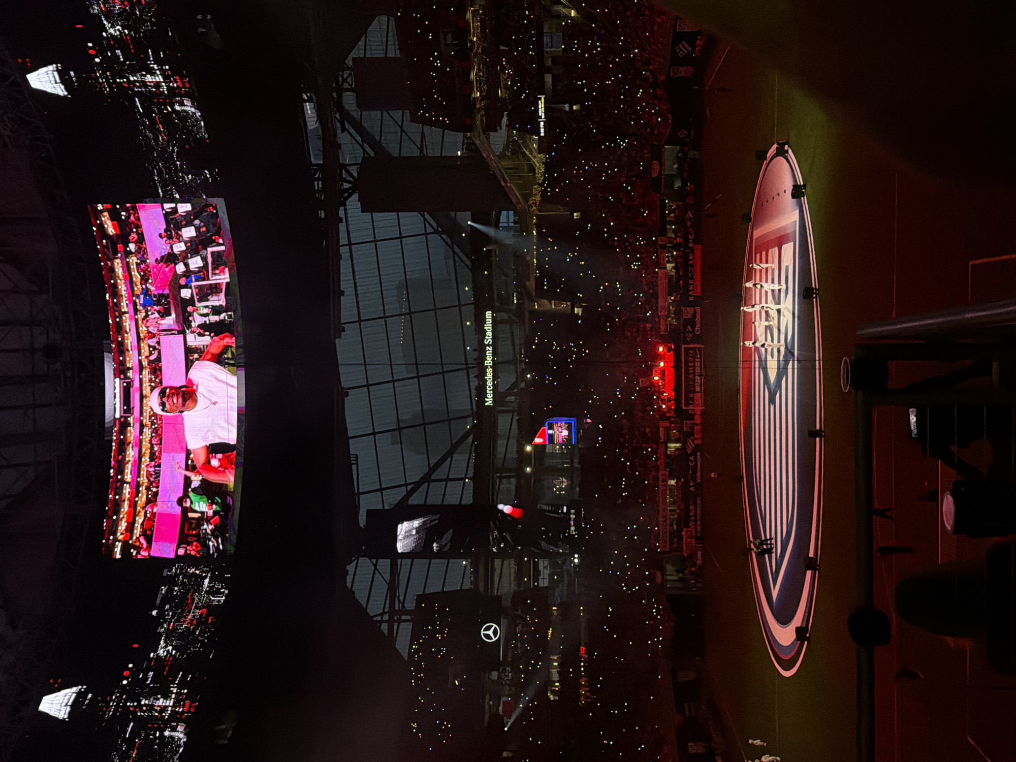 The image shows the interior of the Mercedes-Benz Stadium in Atlanta, Georgia, during an event. The stadium is filled with people, their lights creating a scattered effect in the dark setting. A large screen displays a person on stage. The Atlanta United logo is projected onto the field.