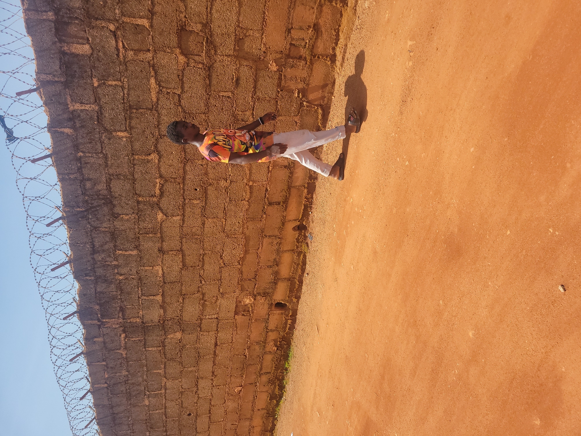 A young man walks along a dusty, orange-colored path next to a tall, rough brick wall topped with barbed wire. He is wearing a vibrantly colored t-shirt with an abstract design and light-colored trousers. He is also wearing dark sandals and has a bracelet on his left wrist. The sun is casting a shadow of the man on the path. The wall is made of roughly textured, rectangular bricks and appears to be a boundary for a property. The sky is clear and blue. This scene is typical of many areas in Jos, Nigeria, where such walls and dusty paths are common.