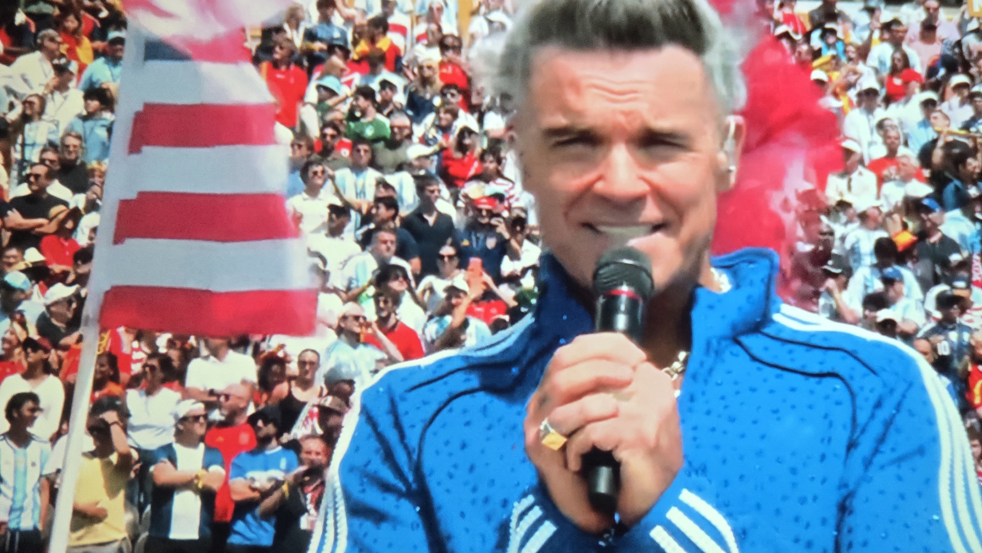The image captures a moment during an outdoor event, likely a sporting match given the presence of a flag with red and white stripes, resembling a national flag, and a large crowd of spectators in the background. A man, prominently featured in the foreground, is holding a microphone and appears to be speaking. He has gray hair and is wearing a bright blue tracksuit with white stripes on the sleeves. He is also wearing a ring on his right hand. The crowd behind him is a mix of men and women, dressed in casual attire, with many wearing shirts in shades of white, blue, and red, possibly team colors. The overall atmosphere suggests a lively public gathering. The location is identified as Kaliningrad, Russia.