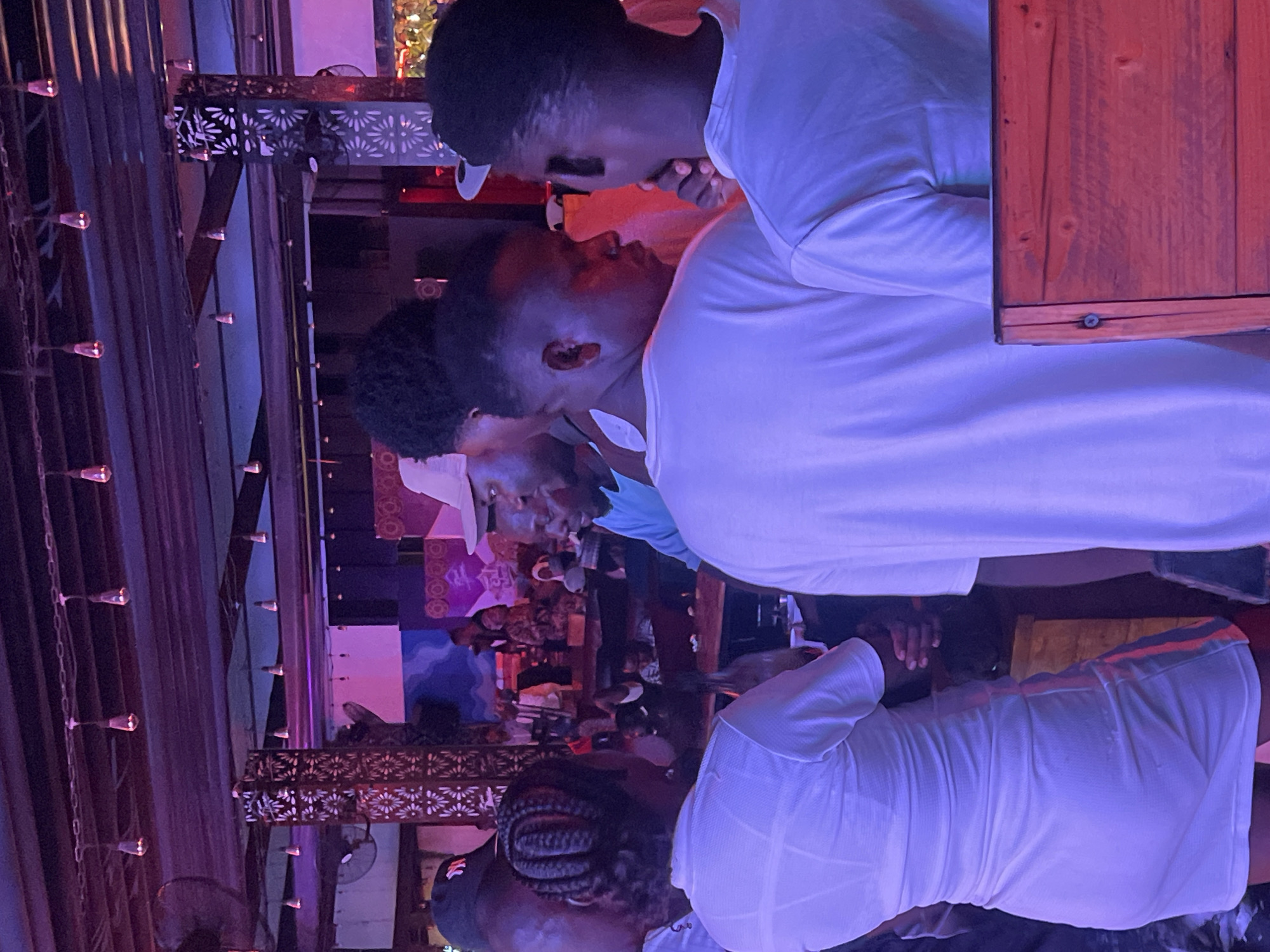 The image depicts a gathering of people indoors, likely in a bar or club in Howayi, Nigeria. The lighting is predominantly blue and purple. Several individuals are seen wearing white clothing, including what appears to be white t-shirts and dresses. Some are standing, while others may be seated in the background. Details like the hairstyles of some individuals, such as braids, are visible. The setting is characterized by decorated metalwork, string lights, and a crowd of people engaged in conversation or socializing.