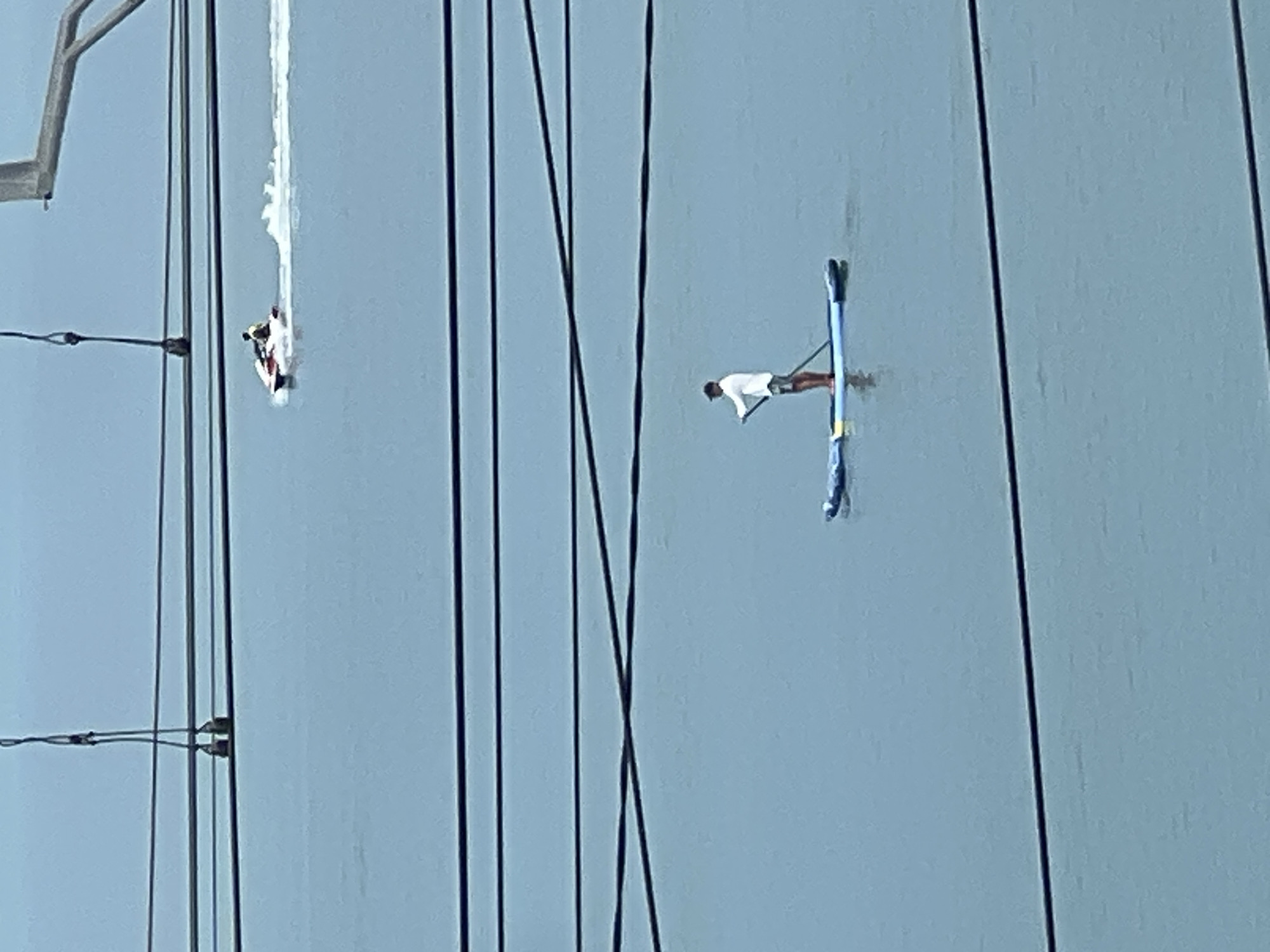 An overhead shot captures a person on a paddleboard in calm, grey water, likely a body of water in Sochi, Russia. The person, wearing a white shirt and dark shorts, stands on a blue and yellow paddleboard, holding a paddle. In the distance, a jet ski speeds across the water, leaving a white wake. Several dark, horizontal lines, possibly power lines or bridge supports, crisscross the frame, creating a layered effect over the water. The sky is a pale, hazy blue, suggesting an overcast or foggy day. The scene appears to be captured from a high vantage point, looking down at the water and activities below.