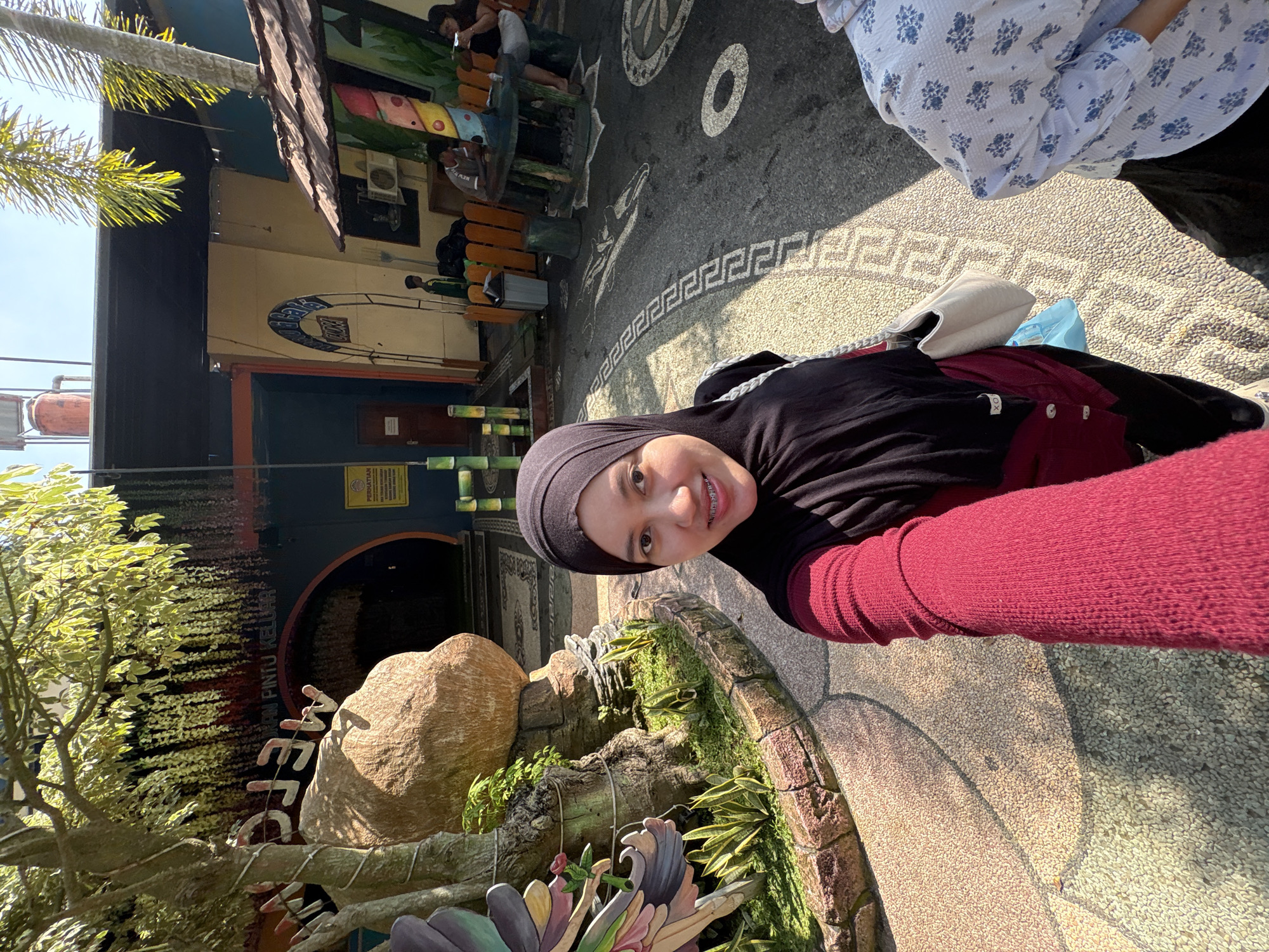 A woman in a hijab and a red sweater is taking a selfie outdoors. She is smiling at the camera, showing off her braces. Behind her, a large rock formation is visible, along with lush greenery and a stone pathway. Further in the background, there's a building with an arched entrance, a sign that reads "PINTU KELUAR," and several people sitting at tables outside. The scene appears to be in a public area, possibly a park or attraction, located in Kebonsari, Indonesia. The ground is paved with small stones and features a decorative pattern. The weather is sunny, casting shadows on the ground.