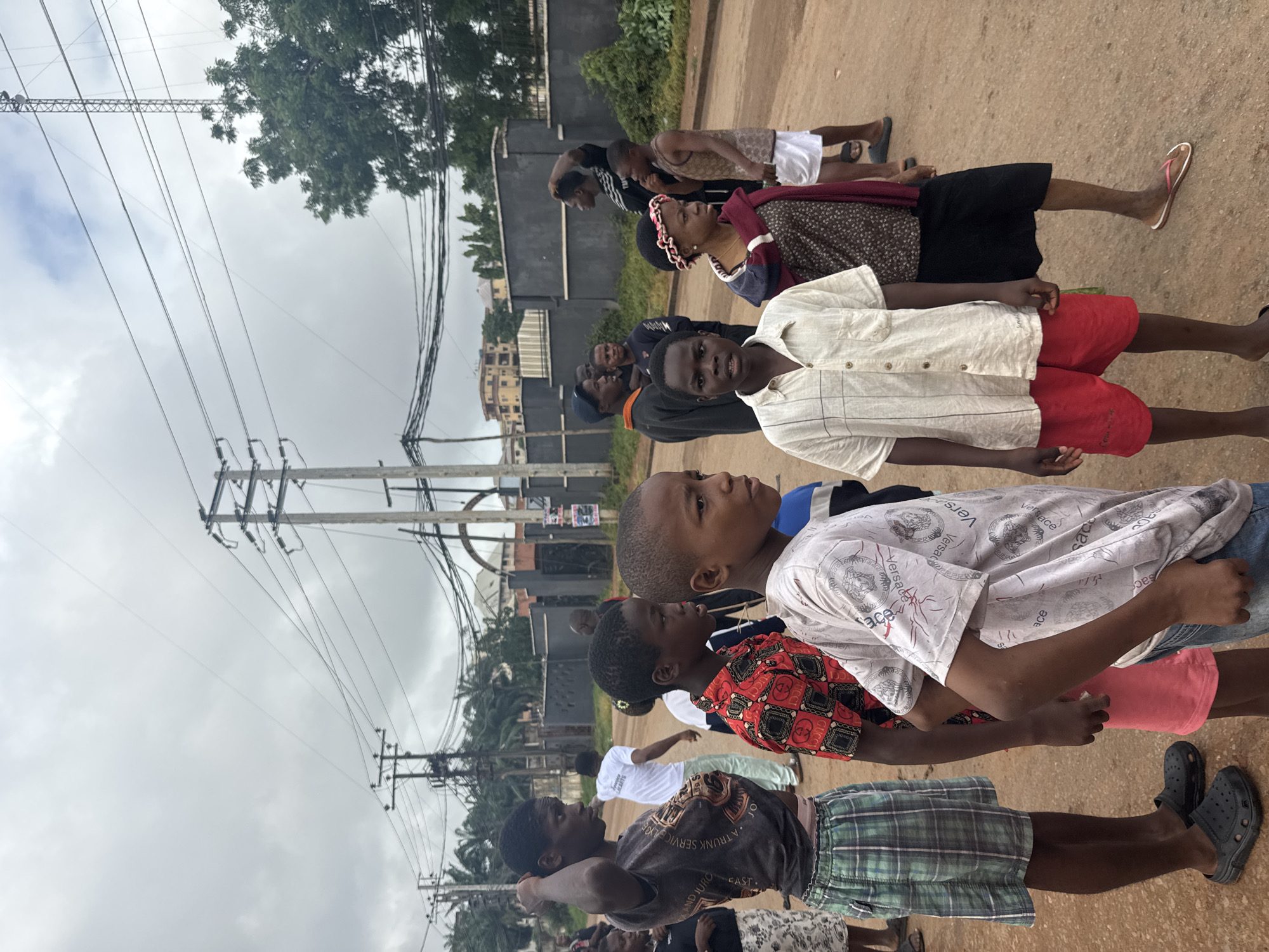 A group of children and young adults stand on a dirt road in Onitsha, Nigeria, under a cloudy sky. The scene is characterized by overhead electrical wires and poles, typical of urban infrastructure. Several people are facing to the right, seemingly looking at something outside the frame. Some individuals wear casual clothing like t-shirts, shorts, and skirts. One person in the foreground is wearing a white patterned shirt and red shorts, while another to their left wears a red shirt with a repeating black pattern. The background shows a dark-colored wall, vegetation, and buildings. The overall atmosphere appears to be a moment captured during an ordinary day in the city.