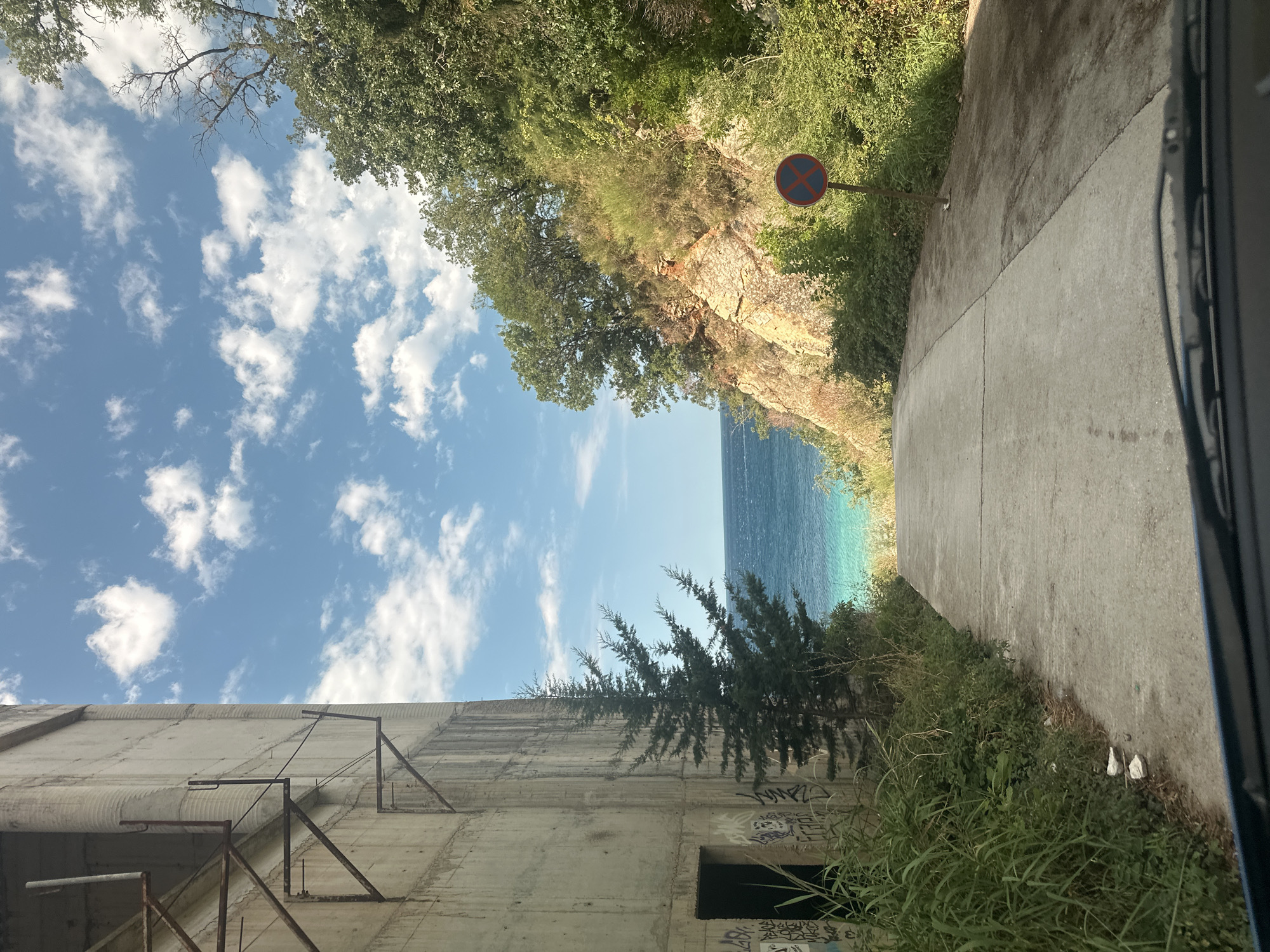 A downward sloping concrete road leads towards a bright turquoise body of water, likely the Adriatic Sea, in Perazića Do, Montenegro. On the left side of the road, a large, weathered concrete structure, possibly an abandoned building or industrial facility, rises with exposed rebar and graffiti. A tall, slender evergreen tree stands in front of the concrete structure. The right side of the road is bordered by a steep, rocky embankment covered in dense green vegetation and trees. A red and blue circular sign with a red cross through it, indicating "no parking" or "no stopping," is visible on the right side of the road. The sky above is a clear blue with scattered white clouds. The image appears to be taken from inside a vehicle, with the car's dashboard and windshield wipers visible in the foreground.