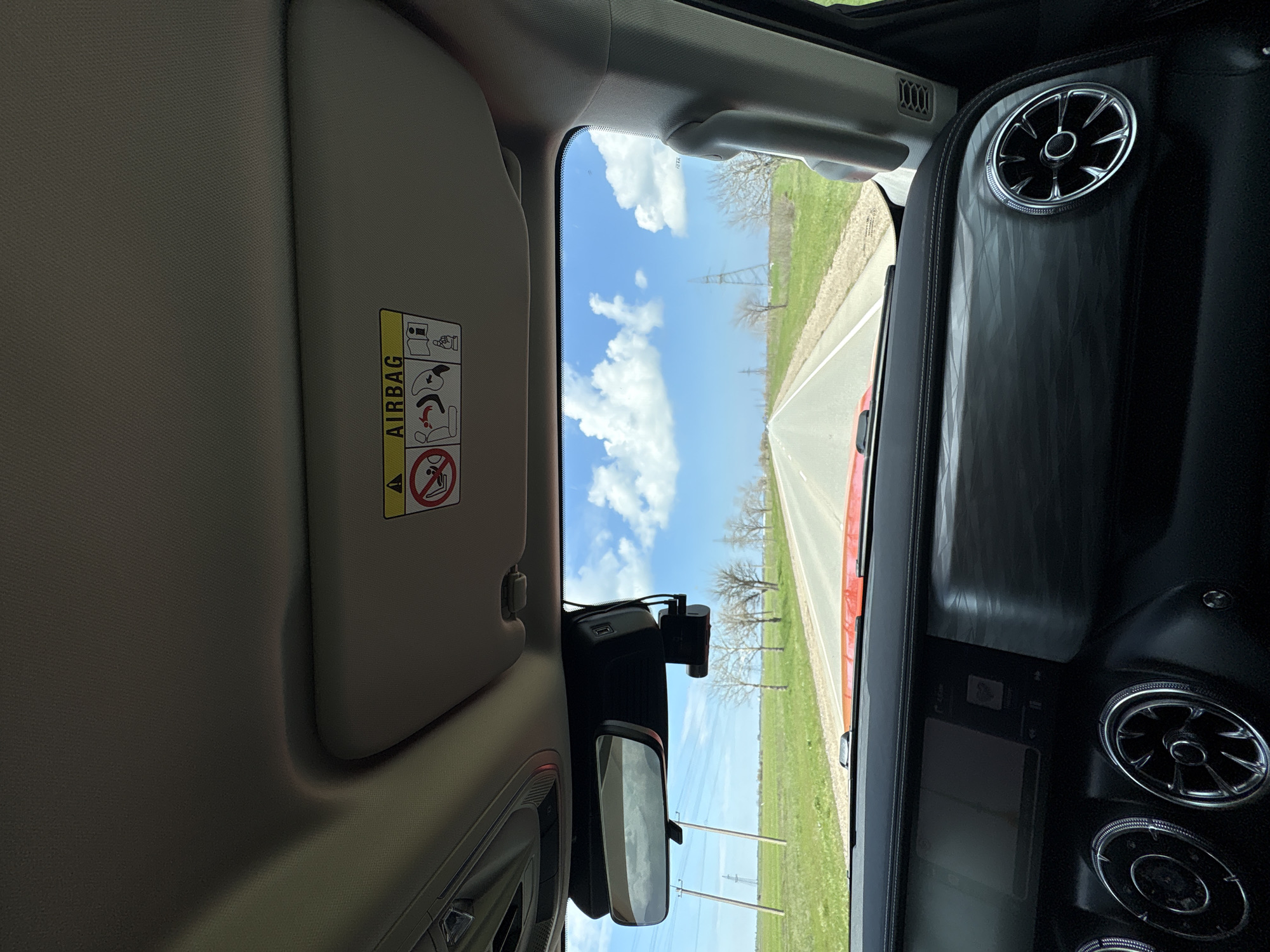 A view from inside a car moving along a straight, seemingly rural road in Николаевское сельское поселение, Ukraine. The sky is blue with scattered white clouds. Green fields border the road, dotted with trees and power lines. The car's interior features a sun visor with an airbag warning label, a rearview mirror with a dashcam mounted, and part of the dashboard with circular vents. The road ahead appears to be clear.