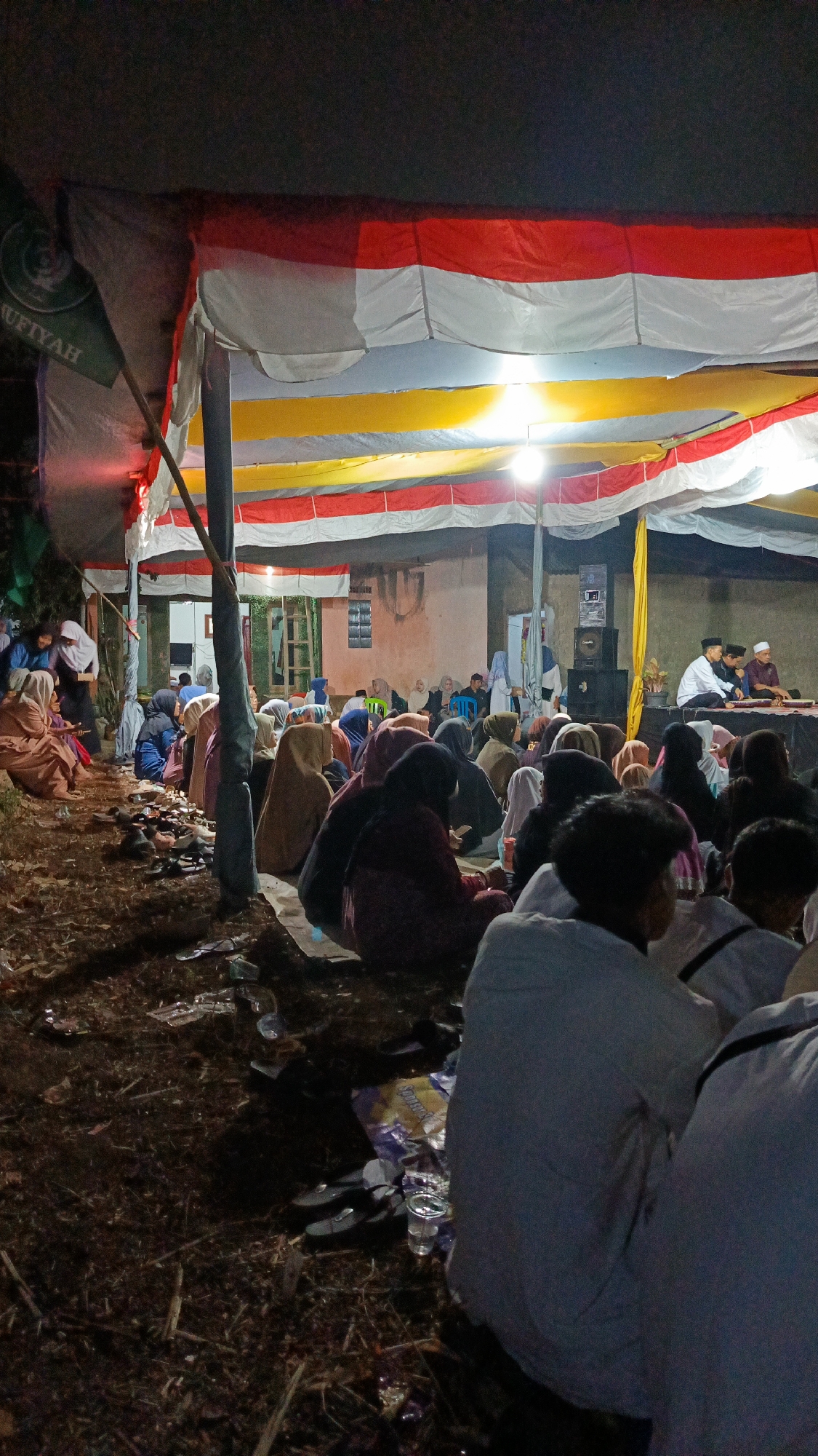 The image depicts an outdoor gathering at night, likely in Margajaya, Indonesia. A large tent covers the area, adorned with red, white, and yellow striped fabric, reminiscent of the Indonesian flag. Several individuals, primarily women wearing headscarves, are seated on the ground, facing a raised platform where three men in white attire and caps are engaged in conversation or presentation. A speaker system is visible behind the men. The ground is covered with dry leaves and scattered debris, including plastic cups and wrappers, suggesting a recent event. Lighting comes from overhead lamps, casting shadows and illuminating the scene. The overall atmosphere appears to be a religious or community event.