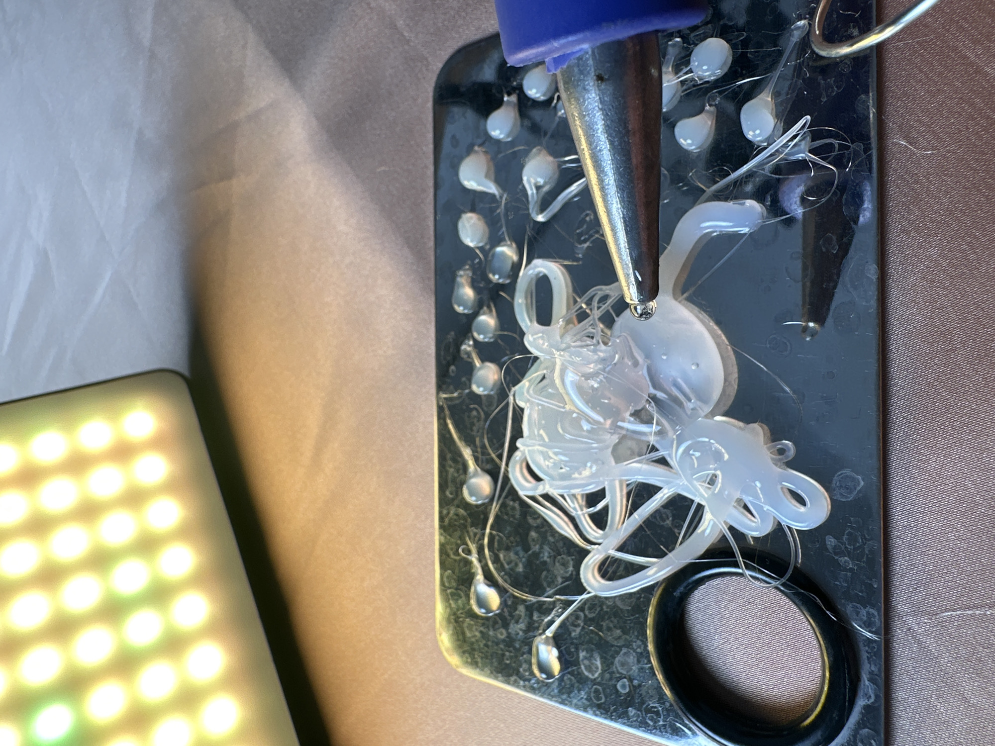 This close-up, high-angle shot captures a hot glue gun in the process of dispensing clear, molten glue onto a dark, reflective surface. The glue gun, featuring a blue plastic top section and a metallic nozzle, is positioned above a significant accumulation of translucent hot glue, which forms a tangled mass along with numerous individual, solidified droplets and fine, stringy wisps. A fresh bead of molten glue hangs from the tip of the nozzle.
The dark surface, possibly a crafting mat or tray, appears slightly worn with visible smudges and traces of dried glue. A thin, metallic wire hook is attached to the upper right side of this surface, and a circular, possibly recessed, black object with a textured, fabric-like interior is visible towards the bottom right.
To the lower left, a bright LED light panel casts a warm, yellowish glow, its grid of small, illuminated square or circular elements clearly visible. The background consists of soft, draped fabrics: a light grey or white material on the left and top, and a brownish-tan fabric on the right and bottom right. The setting appears to be an indoor workspace or crafting area. There is no visible text in the image, and the time of day is not discernible.
