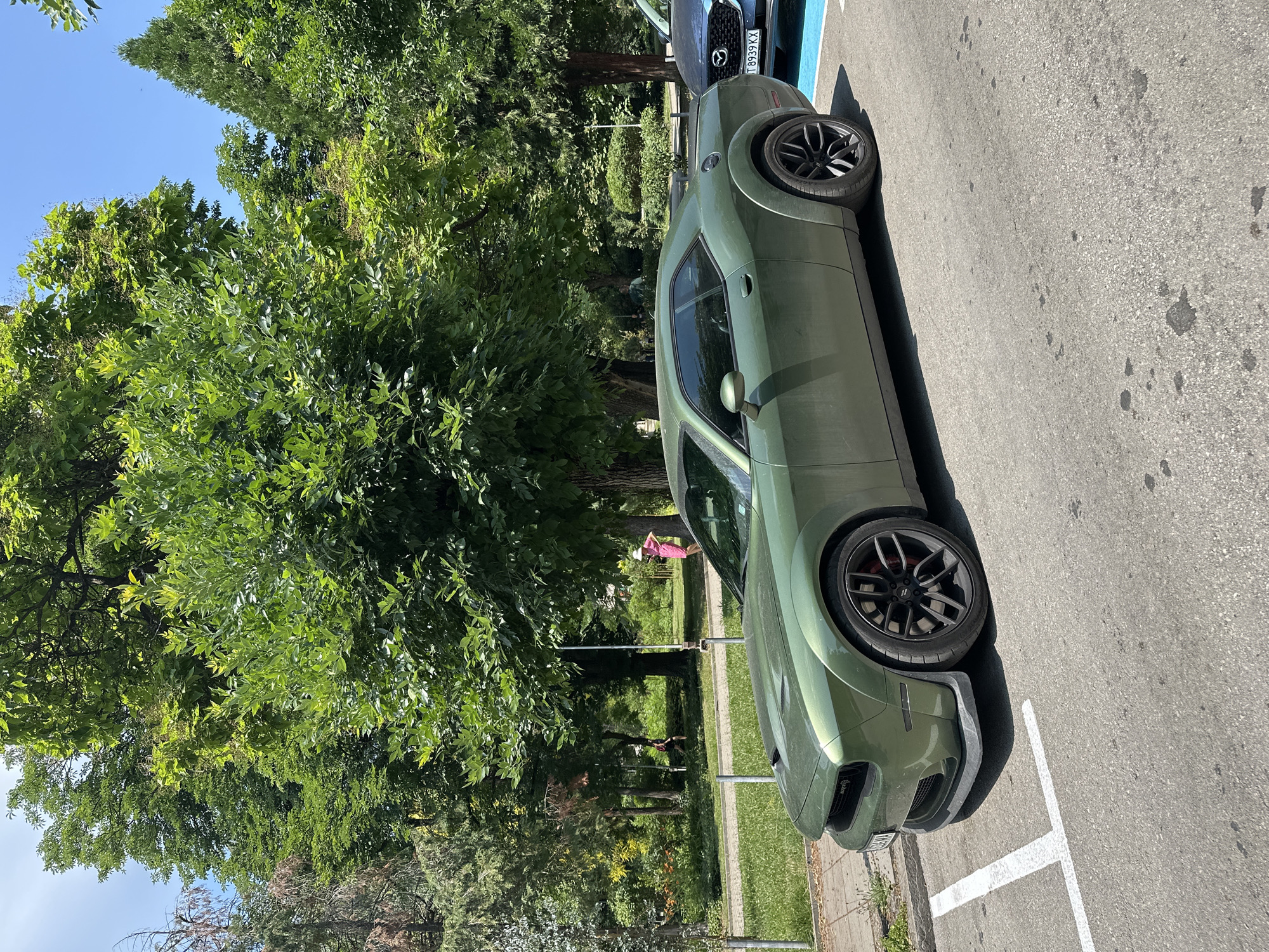 In Veliko Tarnovo, Bulgaria, a modern, military-green Dodge Challenger is parked on an asphalt lot next to a sidewalk and a grassy area lined with trees. The car is angled, showcasing its sleek design and black rims. In the background, a dark blue Mazda CX-5 is parked behind the Challenger, with a Bulgarian license plate reading "T 8939 KX". The scene is bathed in bright sunlight, suggesting a clear day, possibly late morning or early afternoon. A person in a pink dress and white hat is visible walking on the grassy area behind the car, adding a human element to the urban setting. The foliage of lush green trees frames the scene, indicating a pleasant outdoor environment. The overall impression is one of a typical day in a well-maintained park or parking area.
