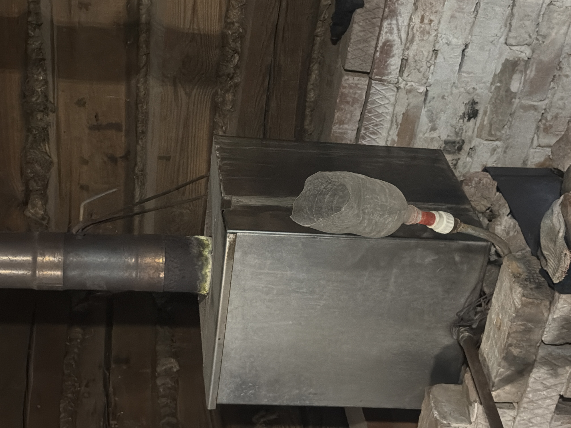 The image shows a close-up, low-angle view of exposed HVAC ductwork and surrounding infrastructure, likely in a basement or crawl space. The primary objects are a large rectangular metal duct, a smaller circular metal pipe running horizontally to the left, and what appears to be a plastic bottle attached to the circular pipe with tape and a hose clamp. The bottle is filled with a gray, lumpy material and has a red and white connector.
The setting is characterized by rough, unfinished materials. The ceiling consists of rough-hewn wooden beams and planks, some of which appear weathered or aged. To the right, a brick wall with a rough, uneven surface is visible, with some decorative or structural elements suggesting an older construction style. There are also some metal conduits and wires visible, indicating the presence of electrical or plumbing systems. The overall impression is one of utilitarian, possibly older, construction, consistent with a service area or utility space.
No people or specific activities are depicted. The lighting is dim, suggesting an indoor environment, possibly without active artificial light sources, or the image was taken with a flash. There is no discernible text in the image. The scene is devoid of any strong emotional cues, presenting a purely observational documentation of the infrastructure. The location context "Аргамак, Russia" provides a geographical reference but no visual cues within the image directly confirm this location.