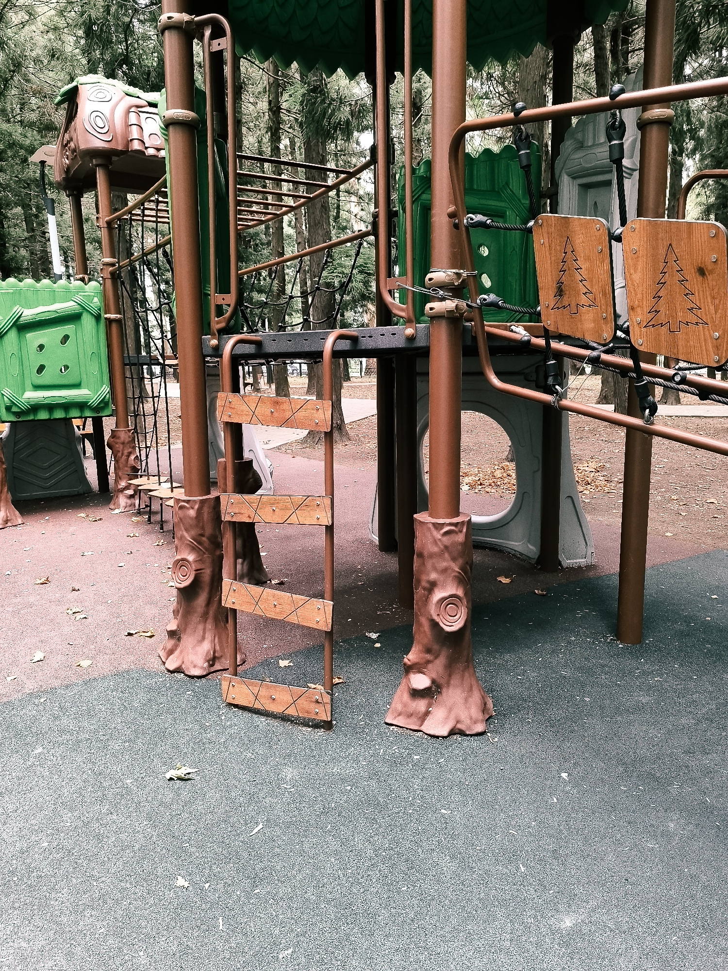 An outdoor playground, possibly located in the city of Batumi, Georgia, features a colorful and multi-level play structure designed to resemble trees. The structure includes brown, tree-trunk-like support columns with sculpted bark details and faux knots. Various play elements are attached to these columns, such as green plastic panels with geometric cutouts, a wooden ladder with zigzagging rungs, and two square wooden panels engraved with tree silhouettes. The playground area is surfaced with a dark, textured material, possibly rubberized safety surfacing, in shades of dark green and red-brown, with scattered autumn leaves on the ground. In the background, tall trees with dense foliage form a natural backdrop. The overall scene depicts a static, unoccupied play area within a natural setting.