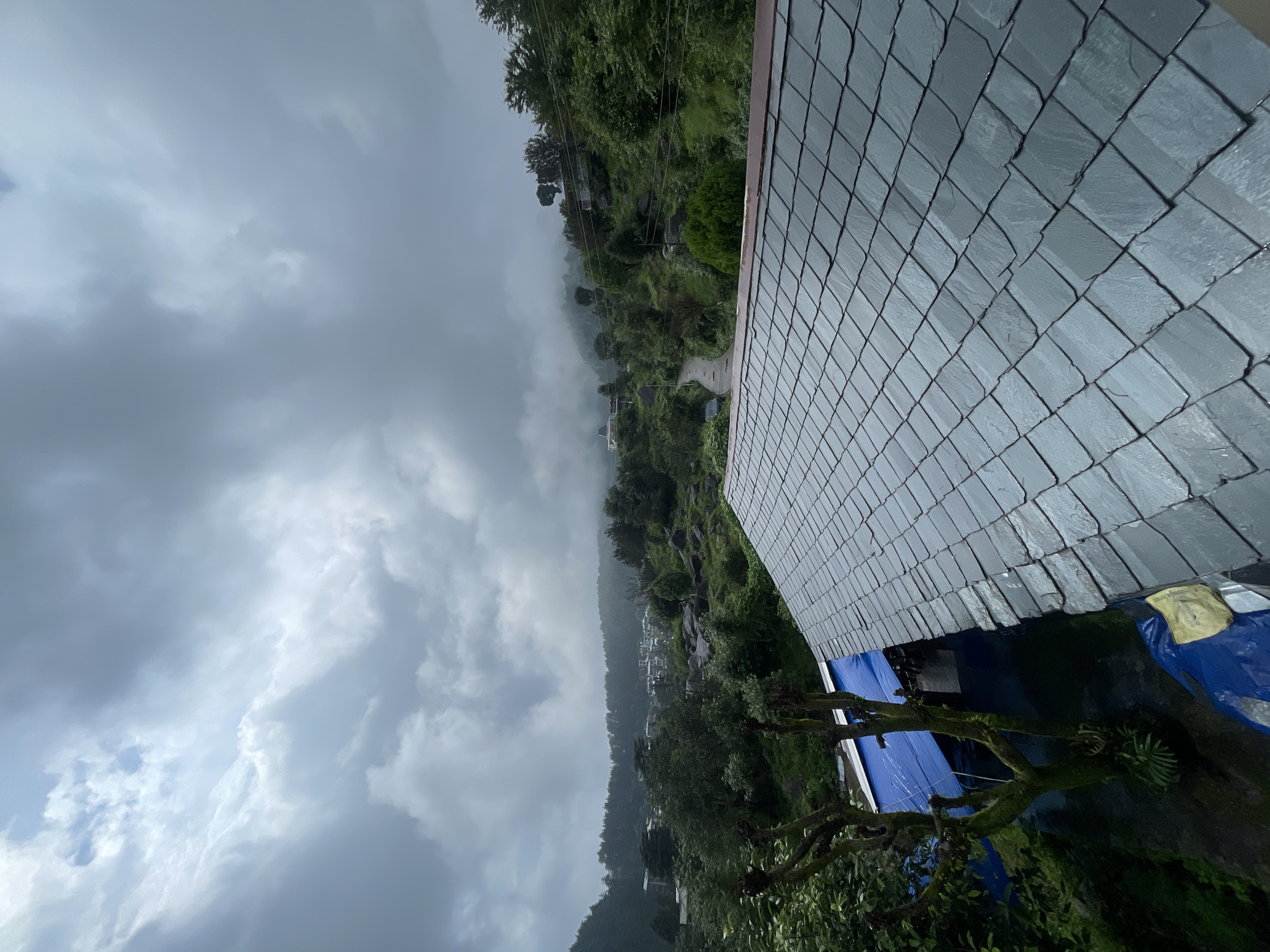 The image depicts a cloudy sky over a village nestled in a valley with lush green hills and trees. A paved road winds through the village, leading towards houses scattered on the slopes. In the foreground, a grey slate roof is visible, with a blue tarp and some vegetation to the left. The scene suggests a rural setting in a mountainous region, possibly during or after rainfall due to the overcast sky and damp appearance of the roof. The location is identified as Haini, India.