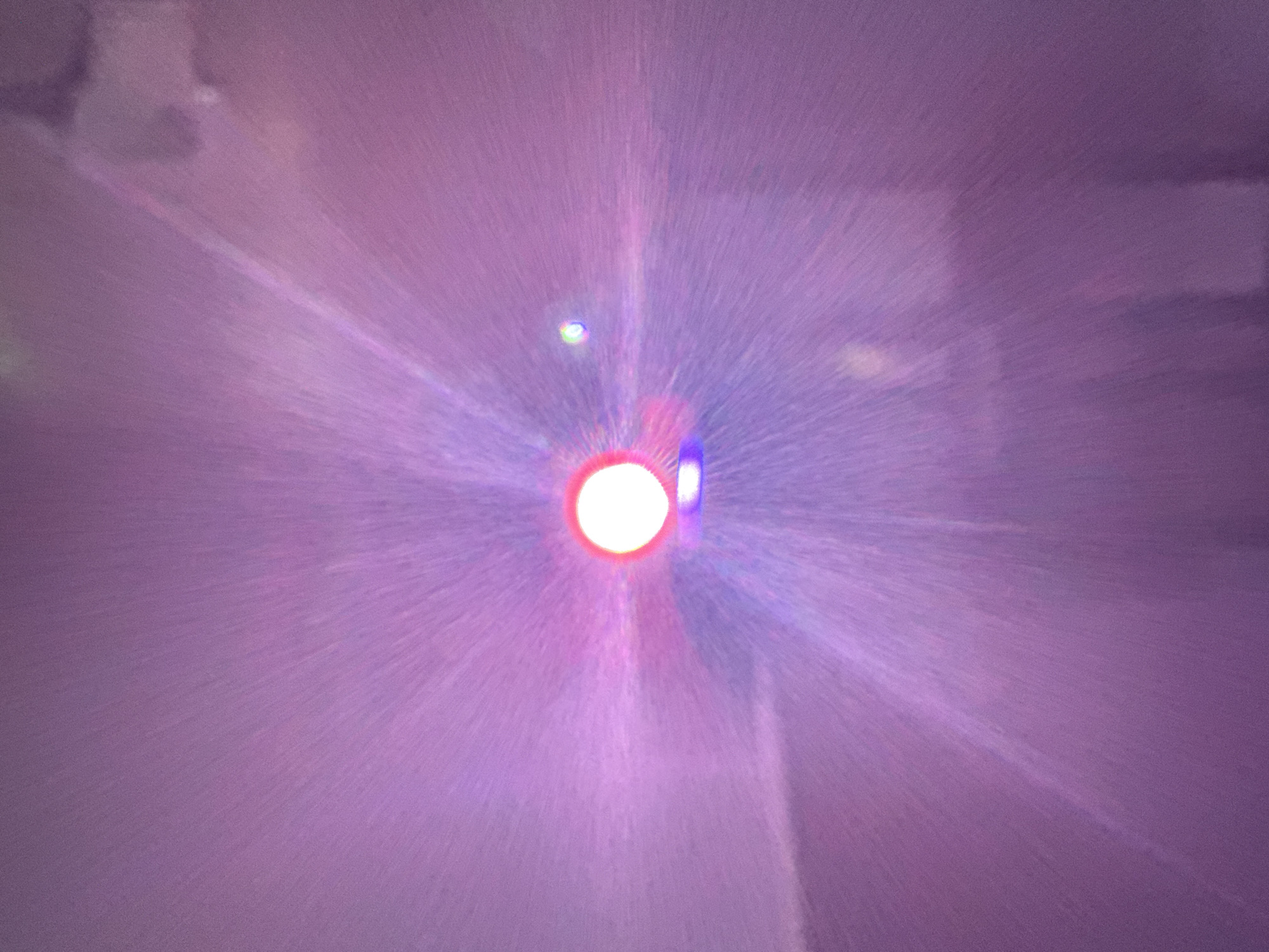 The image depicts a bright, circular light source, likely a lamp or a lens flare, at the center of the frame. Radiating outwards from this light are streaks of purple and pink, creating a starburst or explosion effect. The background is a soft, diffused purple, suggesting a hazy or atmospheric environment. There are no discernible features of a city or specific location within the image. The photograph appears to be abstract, focusing on light and color effects rather than a representational scene. There is no information in the image to connect it to Şalqar, Kazakhstan.
