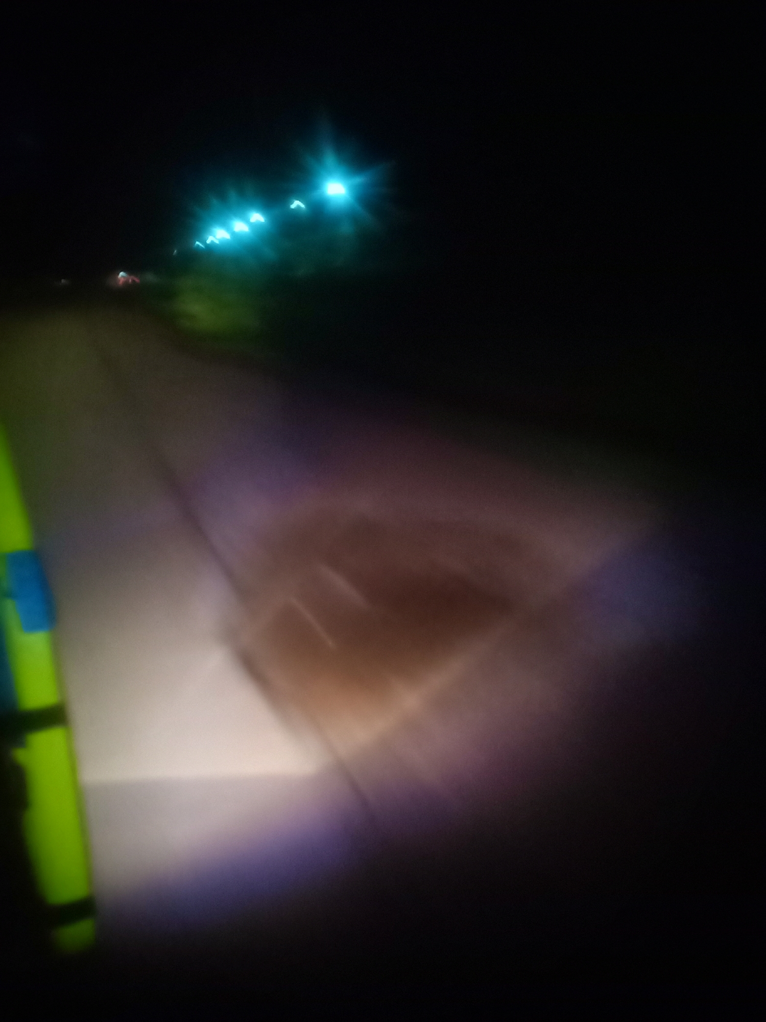 An out-of-focus nighttime image depicts a street or pathway illuminated by artificial lights. On the left side of the frame, a bright green object with black bands is visible, possibly a pole or a sign. Further into the background, a series of blue and white lights are blurred, suggesting they are from vehicles or streetlights in the distance. The ground in the foreground appears to be a light-colored surface, possibly asphalt or concrete, with a faint, rectangular shadow or marking visible on it, illuminated by a warm-toned light source. The overall atmosphere is dark, with a significant portion of the image being black. The location is specified as Unknown, Unknown.