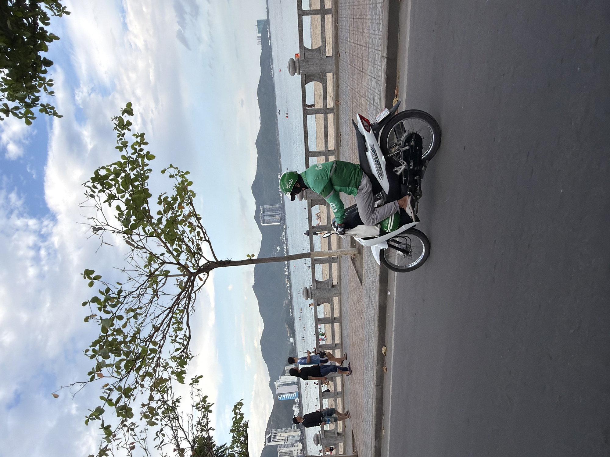 The image depicts a street scene in Nha Trang, Vietnam, likely along the beachfront promenade. The foreground features a grey asphalt road with the rear portion of a white motorcycle in motion, its rear wheel clearly visible. The motorcycle rider is wearing a green jacket with the "Grab" logo and a green helmet, suggesting they are a delivery driver or ride-hailing service operator. The rider is wearing grey pants and white sandals.
Behind the road, a paved sidewalk runs parallel to the beach. Several people are visible walking on the sidewalk. To the left, a group of three men are walking, one in a black long-sleeved shirt and shorts, another in a black t-shirt and jeans, and a third in a blue and white t-shirt and dark shorts. Further along the sidewalk, a woman in a dark dress is walking. The sidewalk is bordered by a decorative concrete wall with circular cutouts, behind which a sandy beach stretches.
The beach is populated with several individuals, some sunbathing and others standing near the water's edge. The ocean is visible, with calm water and a few small boats in the distance. Along the coastline, the city of Nha Trang is visible with numerous high-rise buildings and some mountainous terrain in the background. The sky is partly cloudy with a mix of white clouds and blue sky, indicating daylight. Trees with green leaves are present in the upper portion of the image, framing the scene and adding to the tropical atmosphere.
