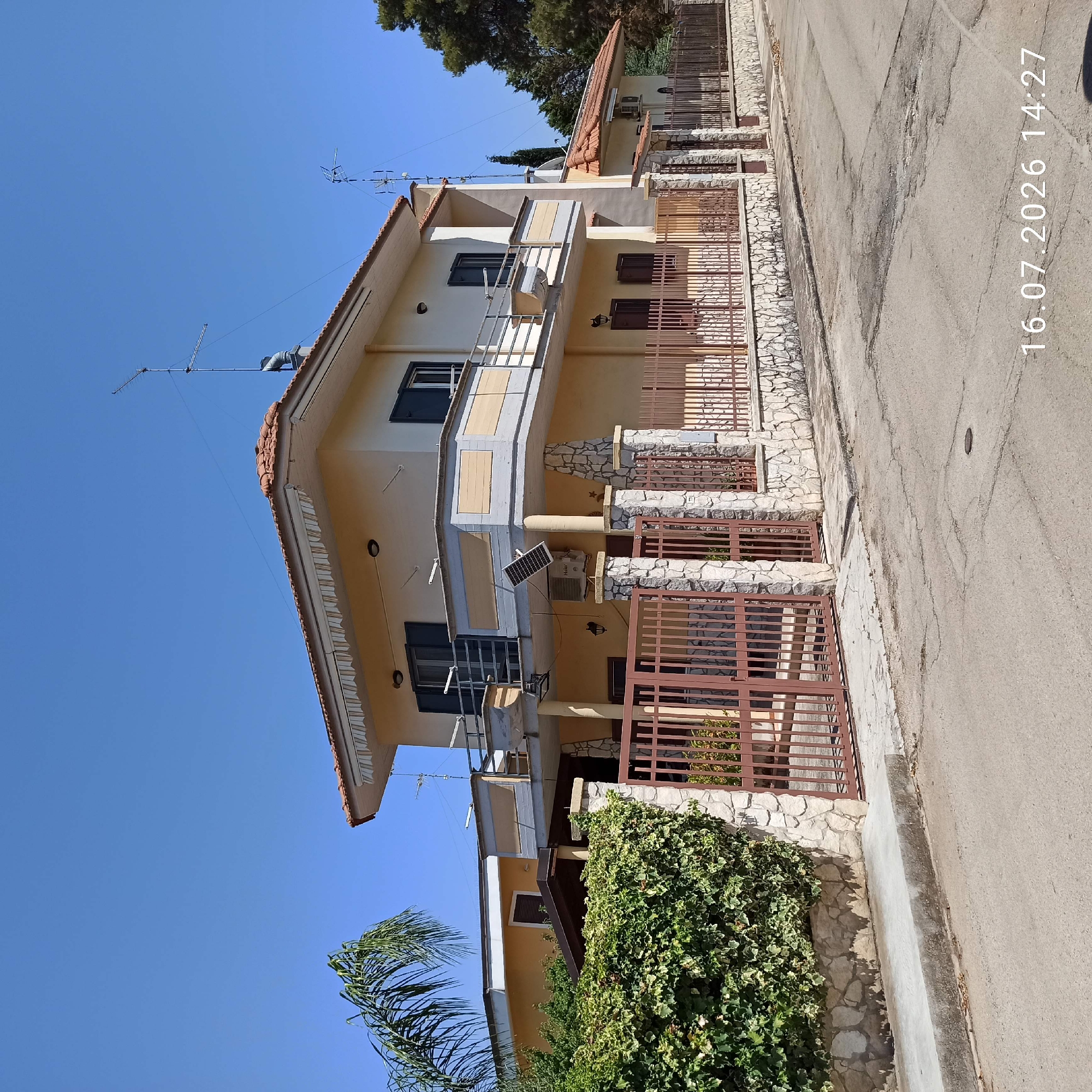 The image shows a two-story house with a light yellow facade, red tile roof, and a balcony on the second floor. The house is surrounded by a stone and metal fence. A lush green hedge is visible along the fence on the left side of the image. A palm tree is also present. The house has several windows with dark shutters. An air conditioning unit and a solar panel are mounted on the exterior wall. The scene appears to be a residential street in Grumo Appula, Italy, with a clear blue sky. The timestamp "16.07.2026 14:27" is overlaid on the bottom right of the image. There are no people visible in the image.