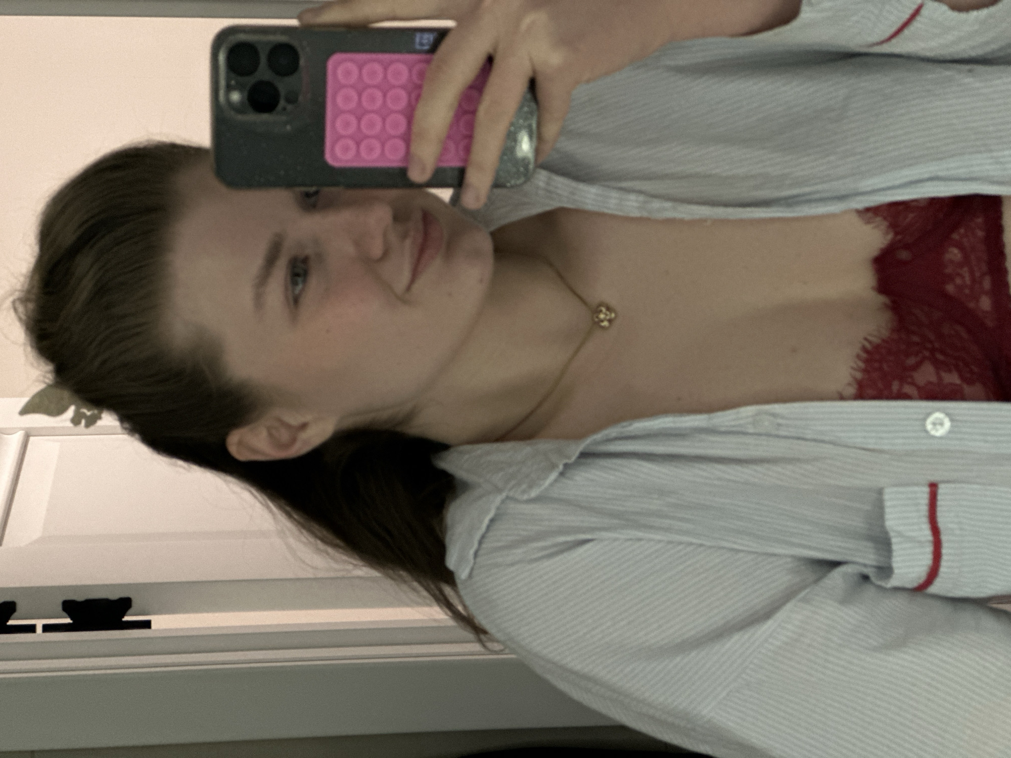 A woman is taking a selfie. She is wearing a light blue striped shirt with red trim on the sleeves. Underneath the shirt, red lace lingerie is visible. She is wearing a gold necklace. Her hair is pulled back, and she is holding a phone with a black case and a pink, patterned grip on the back. The background appears to be an interior, potentially a bedroom or living area, with a door frame visible. There is no indication of Kazan, Russia.