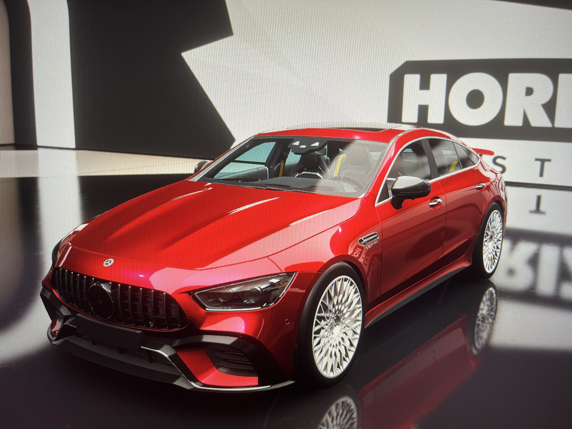 A red Mercedes-AMG GT 63 S sedan is parked on a reflective black surface. The car has custom silver rims. Behind the car, a wall with black and white geometric shapes is visible, with the "HORIZON" logo partially visible on the right side. The setting appears to be a showroom or virtual garage. The city is not known, nor is the country.