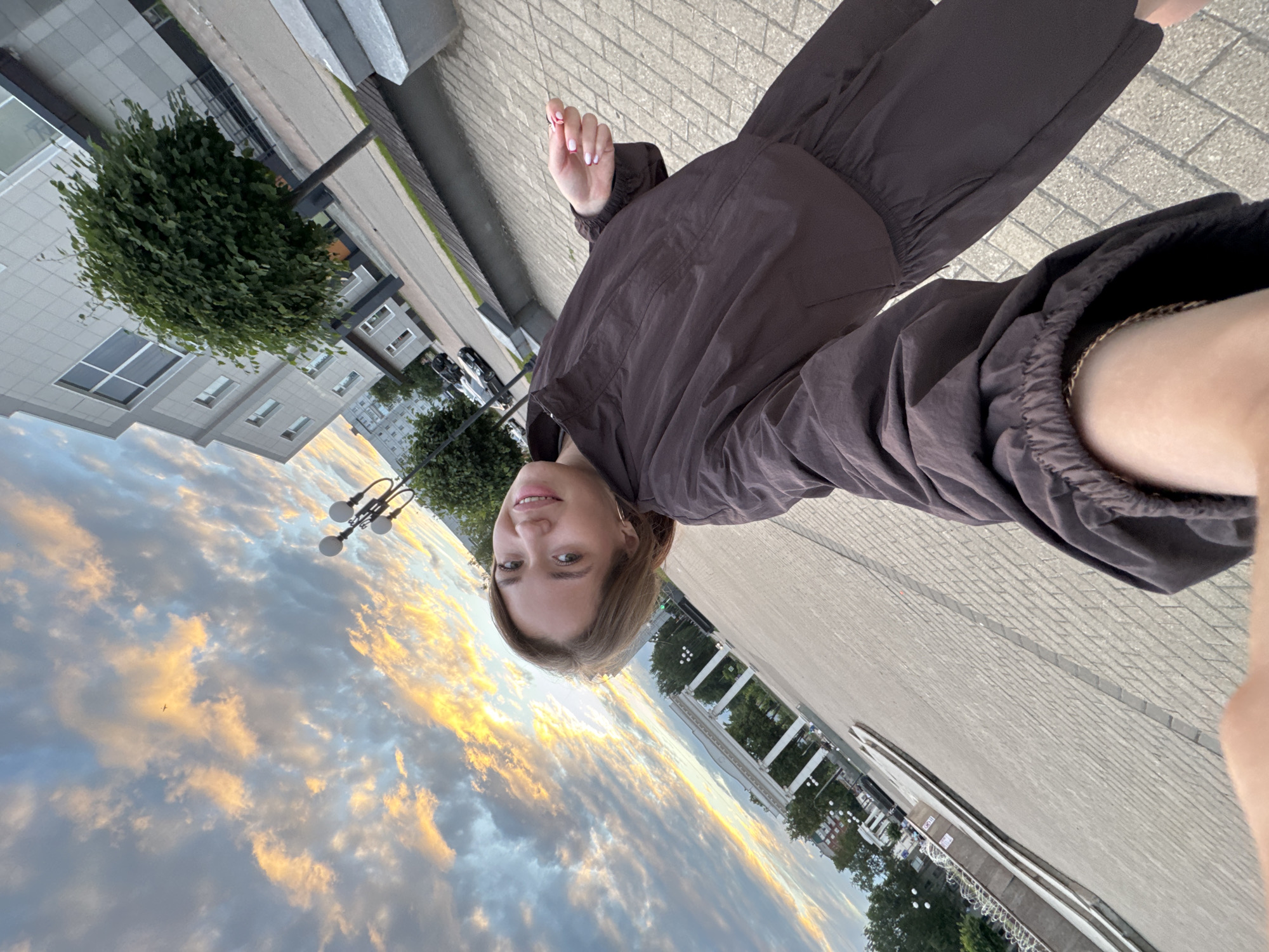 A young woman is taking a selfie in an outdoor urban setting, likely Chaikovsky, Russia, given the architectural style and common scenery of the region. She is wearing a dark brown tracksuit and her right hand, with pink painted nails, is visible near her face, holding what appears to be a small object or accessory. The background features a large, paved square with a distinctive row of white classical-style columns topped with arches, reminiscent of a monument or civic building entrance. Beyond this, there are trees, buildings with modern architecture, streetlights, and a dramatic cloudy sky painted with the warm hues of sunset or sunrise. The ground is paved with rectangular stones, and there are decorative metal fences and benches visible. The overall impression is a moment captured during an evening stroll in a public urban space.
